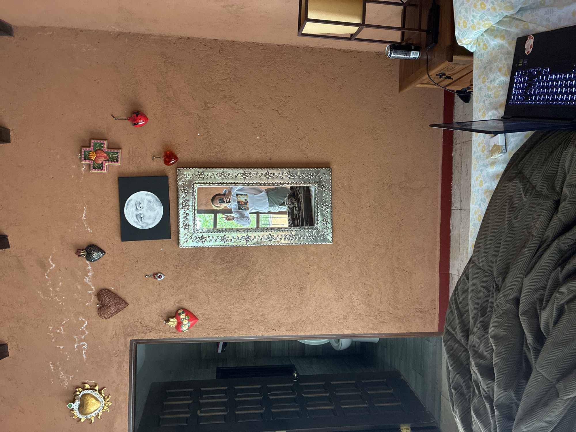The image depicts an interior wall in a residence, likely in Cañada de Madero, Mexico, characterized by textured, brown-orange plaster. A silver-framed mirror hangs on the wall, reflecting a person holding a phone. Various decorative items are affixed to the wall, including metal hearts, a religious cross, and a black square canvas with a white moon. Below, a partially visible dark wooden door with multiple horizontal panels leads into a dimly lit room, revealing a toilet. To the right, a bed with a laptop and a patterned blanket are visible. The overall environment suggests a home interior in Cañada de Madero, Mexico.