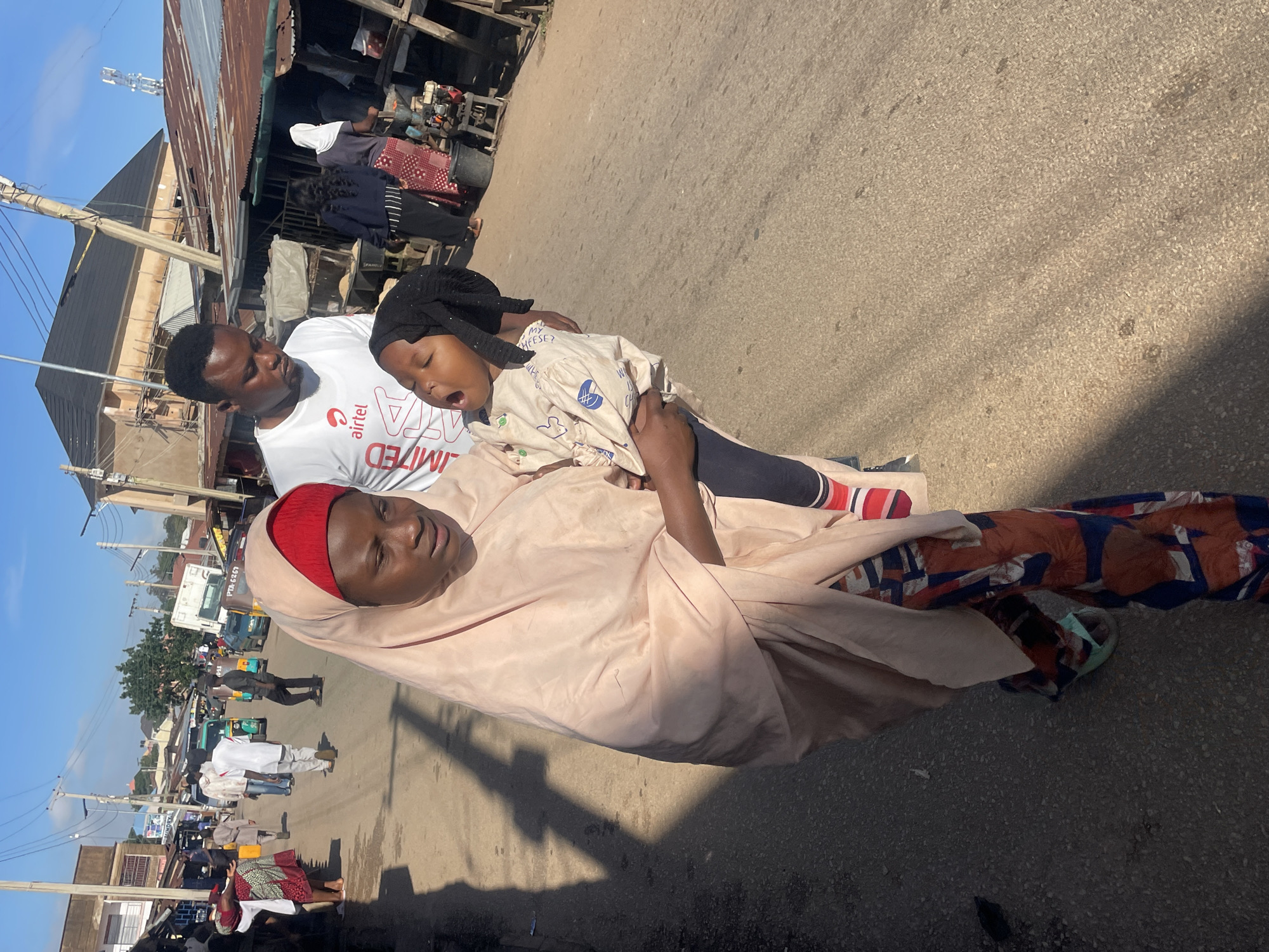 In Jos, Nigeria, on a sun-drenched street lined with market stalls, a woman in a pale pink hijab carries a young child. The child, wearing a black head covering and a patterned shirt with blue designs, is held close, their mouth open as if crying or yawning. Behind them, a man in a white t-shirt with the "airtel" logo walks, looking down. Other people and a few vehicles, including what appears to be a commercial tricycle, populate the busy street. The buildings in the background suggest a bustling urban environment.