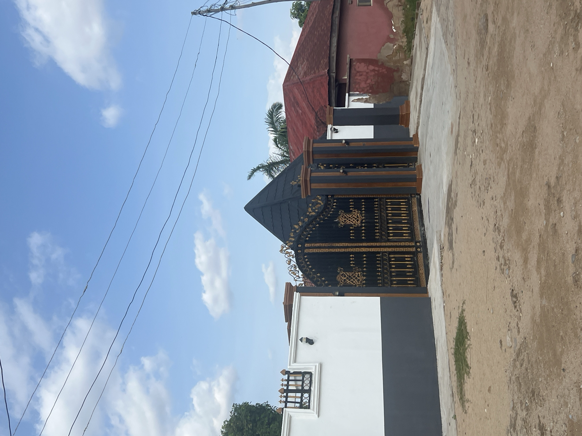 This is a medium shot, eye-level photograph of a large, ornate black and gold gate that serves as the entrance to a property in Jalingo, Nigeria. The gate is intricately designed with a pattern of interlocking shapes and decorative elements. It is flanked by two large, dark grey pillars with wooden accents on top, supporting a conical, dark grey roof that mirrors the design of the gate.
To the left of the gate is a white wall with a dark grey base. A section of this wall features a decorative black metal railing with circular motifs and finials, set within a white rectangular frame. A black outdoor light fixture is mounted on the white wall.
To the right of the gate, a portion of a red-roofed building is visible. The building has weathered, reddish-pink walls with patches of exposed plaster and brick. A small, rectangular window is visible on this building. Behind the gate and to the right, palm fronds are visible, suggesting the presence of trees.
The ground in front of the gate and wall is a mixture of sandy earth and some concrete paving. A small patch of green grass is growing in the sandy area. In the background, a bright blue sky with scattered white clouds is visible. Several black electrical wires are strung across the sky, supported by a utility pole on the far right of the frame. The scene is static and appears to be taken during the daytime.