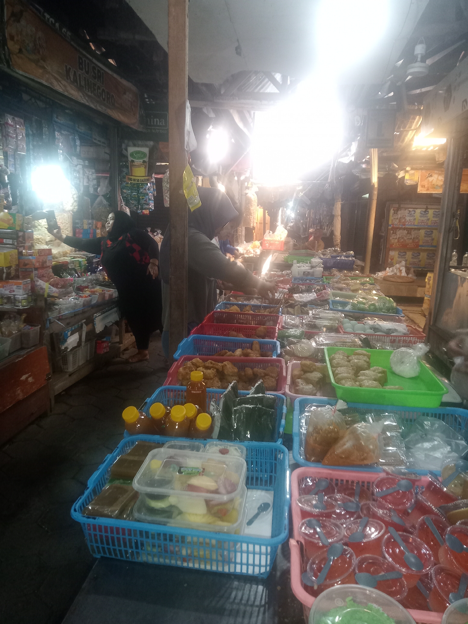 A crowded market stall in Borobudur, Indonesia, is depicted with a bright overhead light illuminating rows of food items. A woman in a black hijab and black garment is taking a selfie with her phone, her arm extended. Next to her, a person in a gray hijab is attending to the stall. The stall displays a variety of food items in plastic containers and baskets, including fried snacks, packaged foods, bottled drinks, and desserts. The background shows shelves stocked with more goods, suggesting a busy commercial environment within the market.