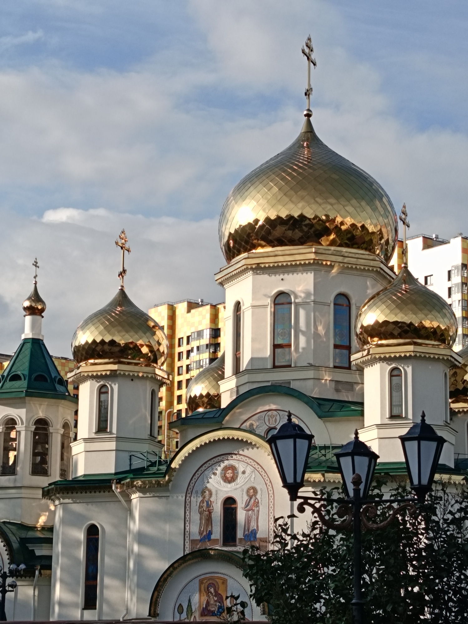 This is a medium shot, taken from a slightly low angle, of an Orthodox church with multiple gold-domed cupolas. The main cupola in the center of the image is large and highly reflective, catching the sunlight. To its left, a smaller green-roofed tower with a gold cupola is visible, and to its right, another smaller gold-domed cupola. The church itself is white with decorative arches and stained-glass windows. Above the entrance, there are mosaics depicting religious figures.
Behind the church, several modern apartment buildings are visible, one with a distinctive yellow and orange facade. The sky is partially cloudy with patches of blue. The lighting suggests it is either late afternoon or early morning, as the sun is casting long shadows and highlighting the golden domes. The overall mood is one of serene religious architecture juxtaposed with modern urban development. Three black lampposts with ornate detailing stand in the foreground, partially obscuring some of the lower portion of the church and lush green foliage. No visible text is present in the image. The setting is identifiable as Saint Petersburg, Russia, given the architectural style of the church and the surrounding environment.
