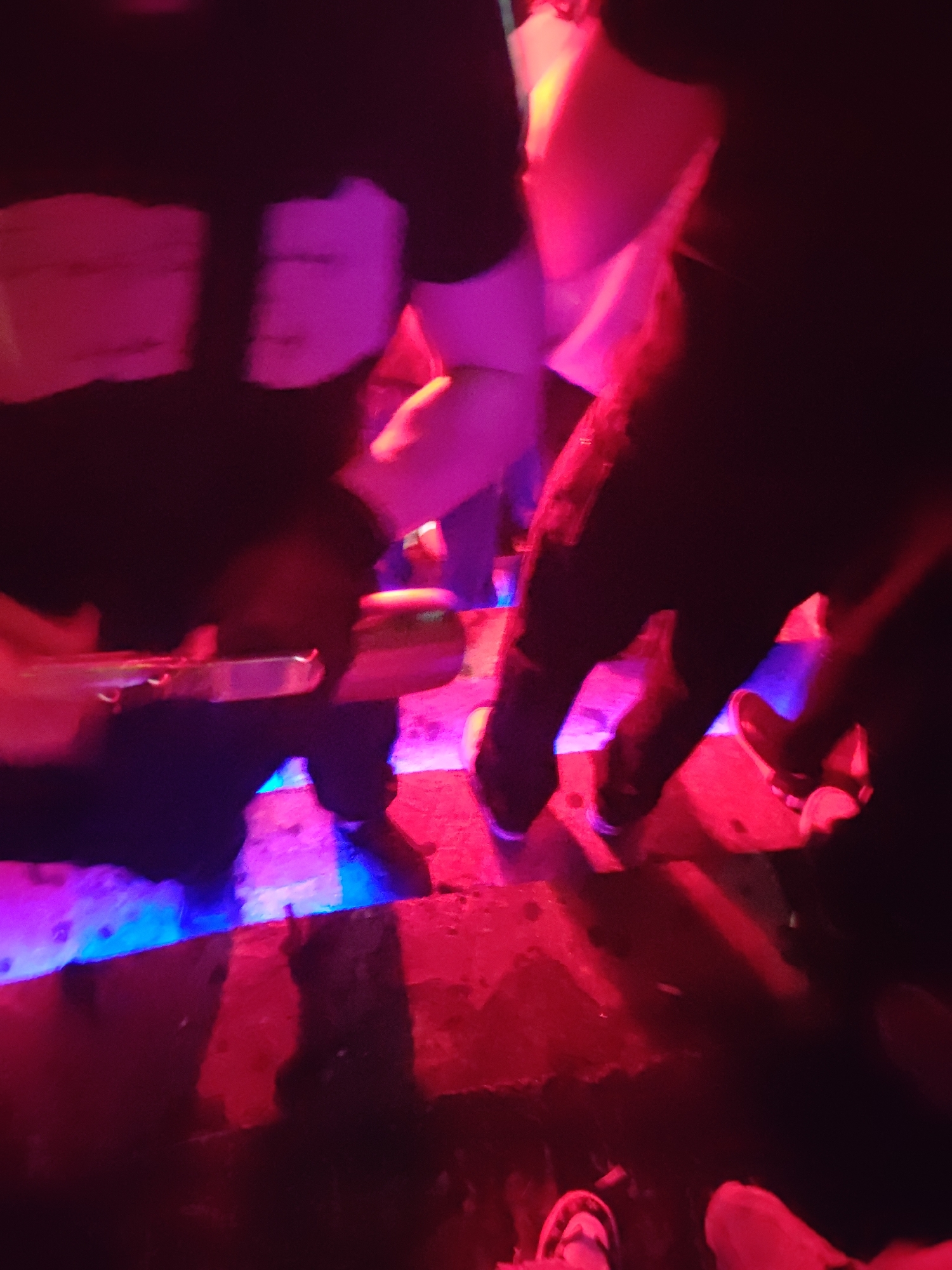 The image depicts an interior scene within an entertainment venue in Varna, Bulgaria. The environment is characterized by low light, intensely illuminated by artificial red and blue sources. A horizontal blue light strip is visible at floor level, with red light saturating the surrounding areas and casting strong shadows.
Multiple individuals are present, their forms partially obscured by darkness and motion blur. A central figure, dressed in dark trousers and a dark long-sleeved top over a lighter garment, is holding a reflective, cylindrical object horizontally with both hands. The object, appearing metallic or glass, possibly a bottle or a bar tool, reflects the ambient red and blue light. The lower limbs and feet of several other individuals are visible on a dark, textured floor, some illuminated by the blue light strip. Their footwear includes light-colored casual shoes. The scene conveys a dynamic and crowded atmosphere consistent with a nightclub or bar setting.