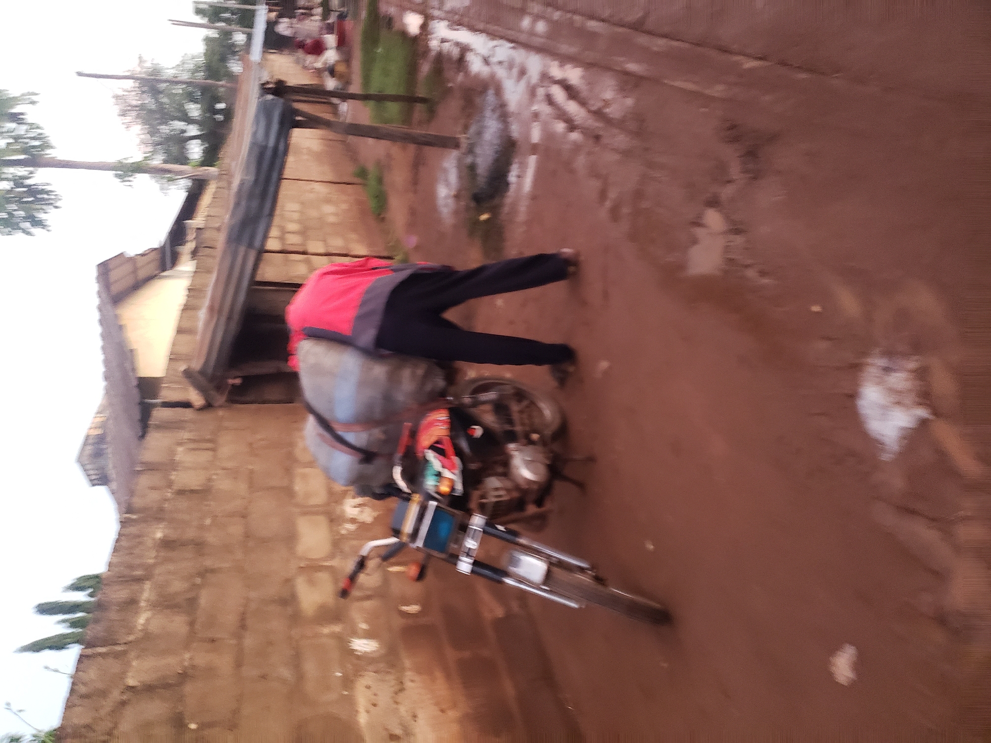 In Kakuri Gwari, Nigeria, a person in a red and grey jacket and black pants is standing next to a motorcycle loaded with a large bundle of grey material, possibly merchandise or personal belongings. The motorcycle is parked on a dirt road that appears wet and muddy, with puddles of water visible. Behind the person and motorcycle is a brick wall, partially obscured by a corrugated metal roof structure that extends over a small outdoor area. A few trees are present in the background, and the sky is overcast, suggesting either early morning or late afternoon lighting. The overall scene depicts a moment of transit or preparation for travel, with the motorcycle serving as the mode of transport for the loaded bundle.