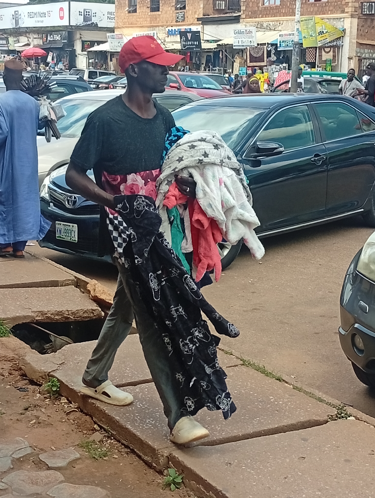 In Jos, Nigeria, a man walks down a sidewalk carrying an armful of clothes. He wears a red baseball cap, a dark t-shirt with a floral pattern on the lower part, and grey pants. His footwear consists of white slide sandals. He is walking past parked cars, including a dark sedan with a license plate that reads "KWL-993CX." The background shows shops and buildings typical of a city street. The ground is paved with concrete, and a drainage ditch is visible along the edge of the sidewalk. The man's attire and the surrounding urban environment suggest a scene of daily life in the city.