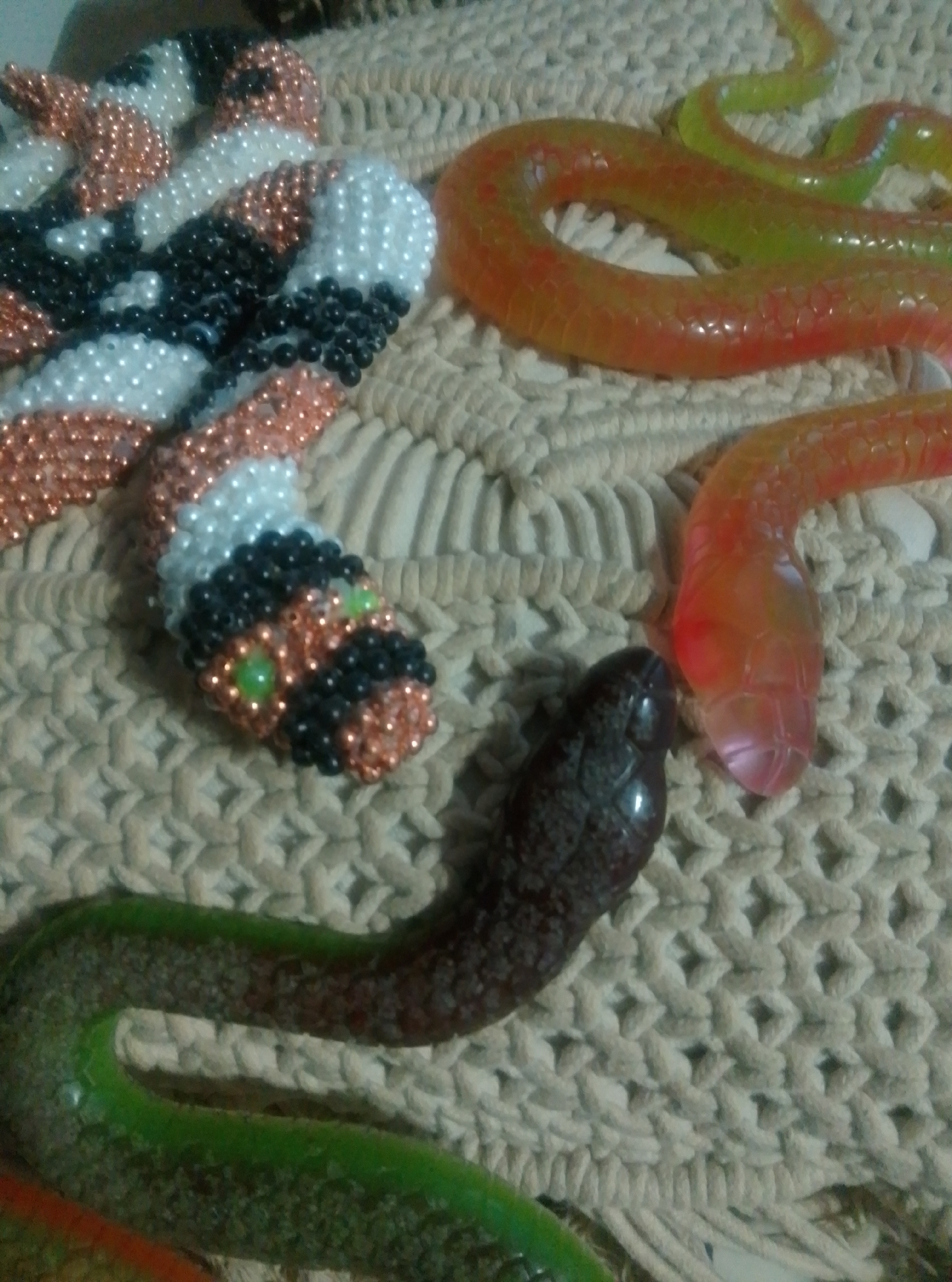 The image displays a collection of toy snakes arranged on a woven surface, likely a rug or placemat. There are at least three distinct toy snakes visible. One snake, in the foreground and to the left, appears to be a beaded craft, possibly a decorative item. It has a head with green bead eyes and is colored with white, black, and copper beads. To its right, a translucent orange and yellow snake coils across the woven background. In the lower right quadrant, another translucent snake, this one colored red and orange, has its head close to the beaded snake's body. A fourth snake, predominantly green with a dark brown patterned body, curves across the bottom of the image. The overall scene is a still life arrangement of these snake toys. There is no indication of people, text, time of day, weather, or specific location cues beyond the general setting of a surface with a woven texture.
