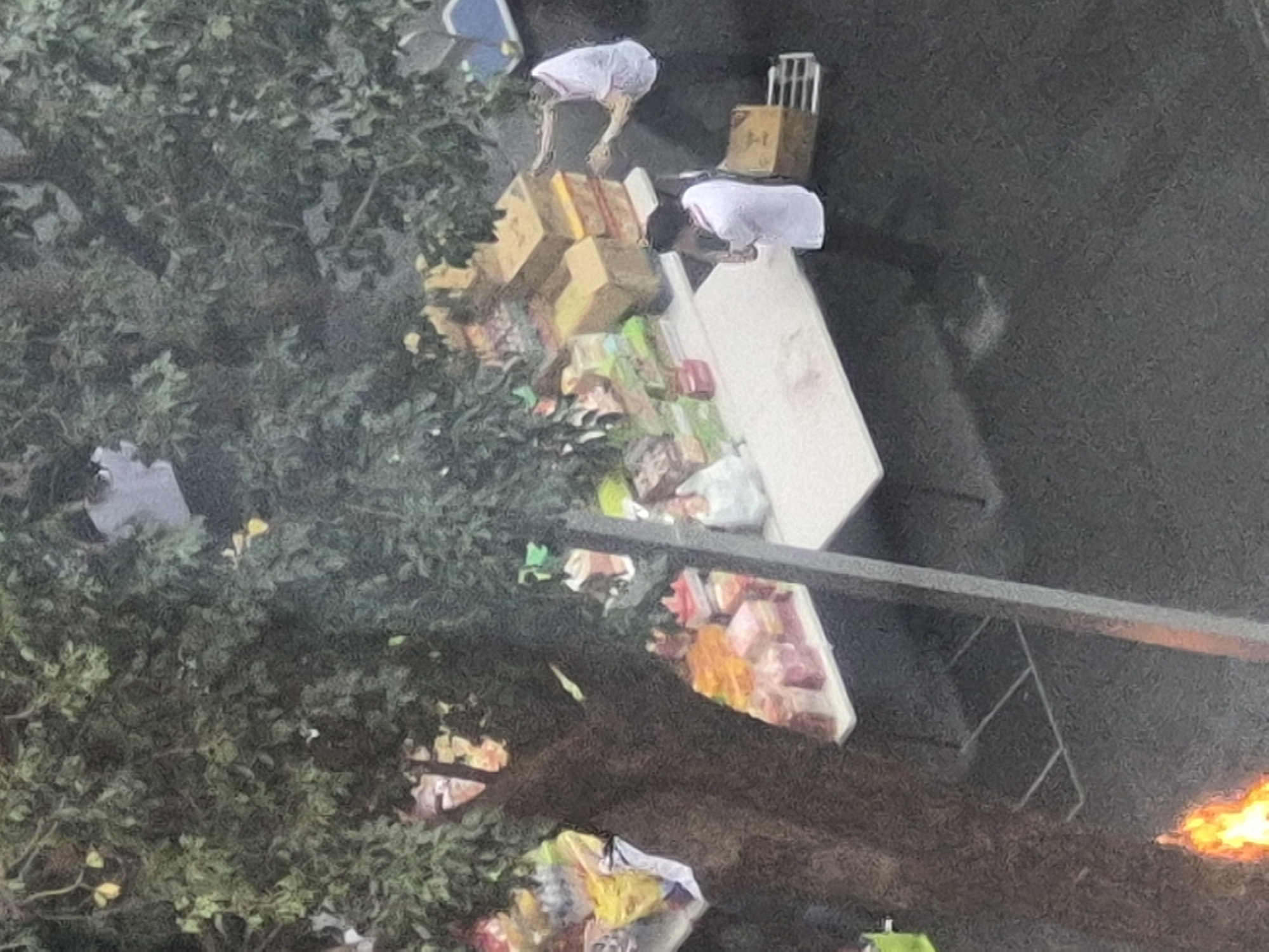 The image depicts an outdoor scene, possibly a street market or a temporary stall, under the canopy of a large tree. On a white table, there are numerous items, including stacked cardboard boxes, what appear to be gift-wrapped packages in various colors (pink, orange, yellow), and some plastic-wrapped goods. Two individuals are present. One person, wearing a white shirt, is standing behind the table and interacting with the items. Another person, also in a white shirt, is standing in front of the table, holding what seems to be a camera or phone, and facing the table. In the background, more items are visible, including bags and possibly more stalls or structures. To the left of the frame, there is a bright orange flame, suggesting a fire or a decorative element. The ground appears to be paved. The overall impression is of a vendor or a public event taking place in a city setting.