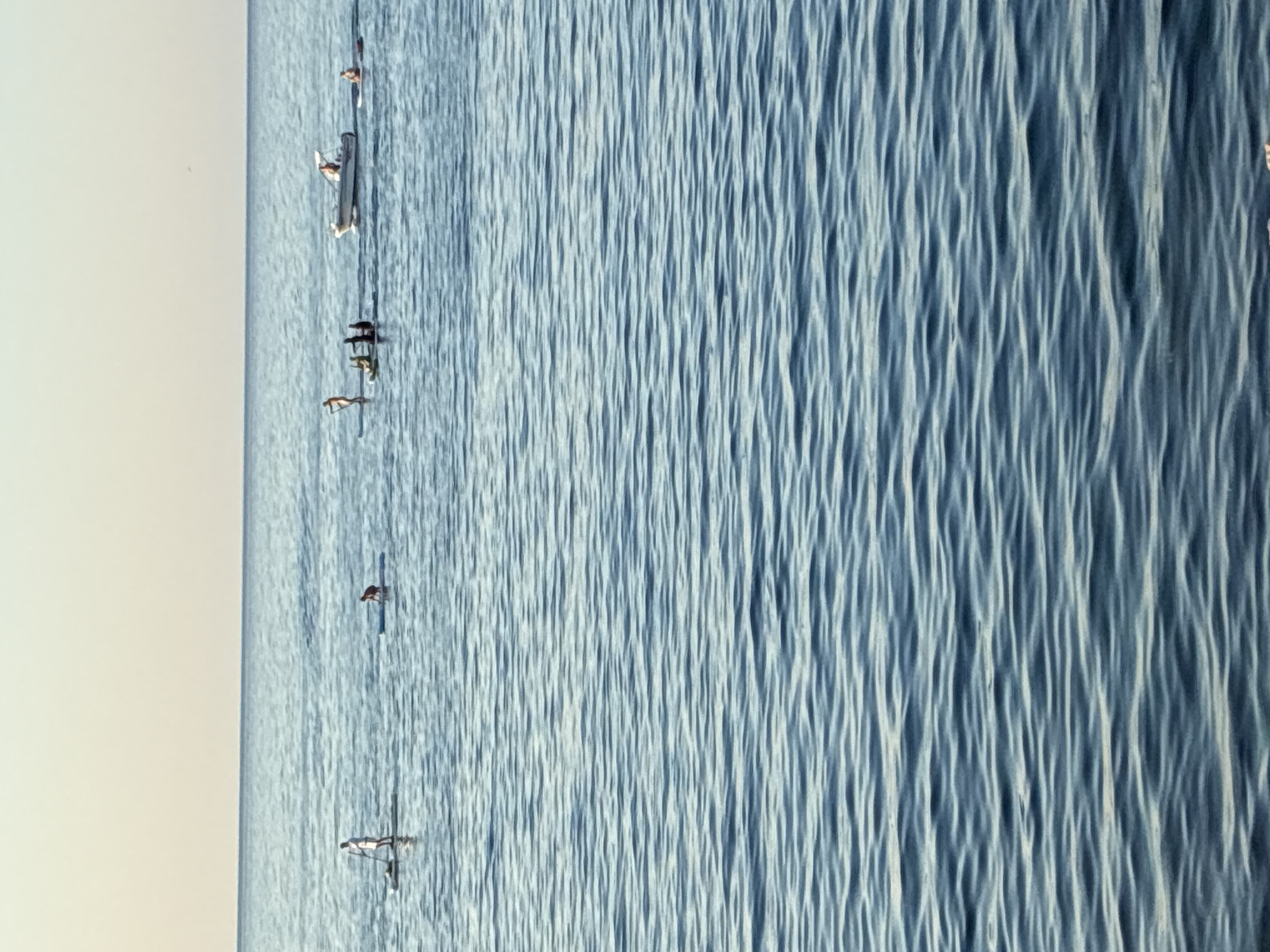 A wide shot captures a calm, blue ocean under a pale sky, with a clear horizon line. Several individuals are engaged in water activities. On the left, a person stands on a paddleboard. To their right, another person kneels on a paddleboard. Further to the right, a group of three people are on a paddleboard, with one in front and two behind. Beyond them, a white motorboat with two visible occupants travels across the water. To the right of the boat, a person sits on a paddleboard. The water has gentle ripples throughout. The scene appears to be set in Alanya, Turkey, given the context provided, likely along its coastline.