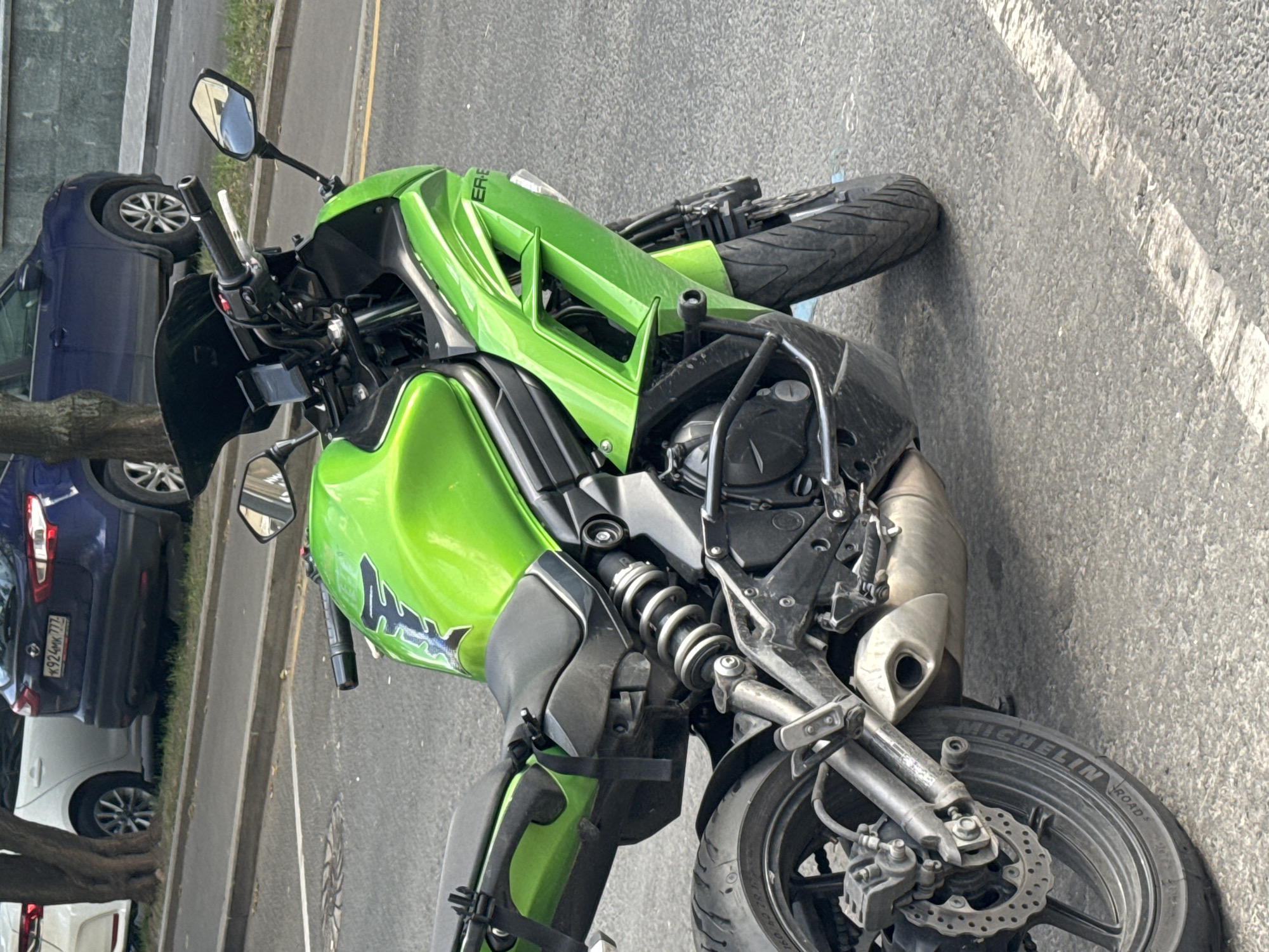 A bright green Kawasaki ER-6f motorcycle is parked on a paved city street, likely in Moscow, Russia, based on the vehicle license plates visible in the background. The motorcycle is positioned at an angle, with its front wheel slightly turned. The street appears to be in a residential or commercial area, with a sidewalk and several parked cars in the background. One of the cars, a dark blue Nissan Qashqai, has a Russian license plate that reads "K924MK 777". The motorcycle has a black windscreen, black handlebars, and a black seat. The rear of the motorcycle features a silver exhaust pipe and a black rear fender. The surrounding environment is bathed in daylight, and the asphalt road surface shows signs of wear and tear, with white markings indicating a lane divider. The scene is static, with no people actively engaged with the motorcycle or in the immediate vicinity.