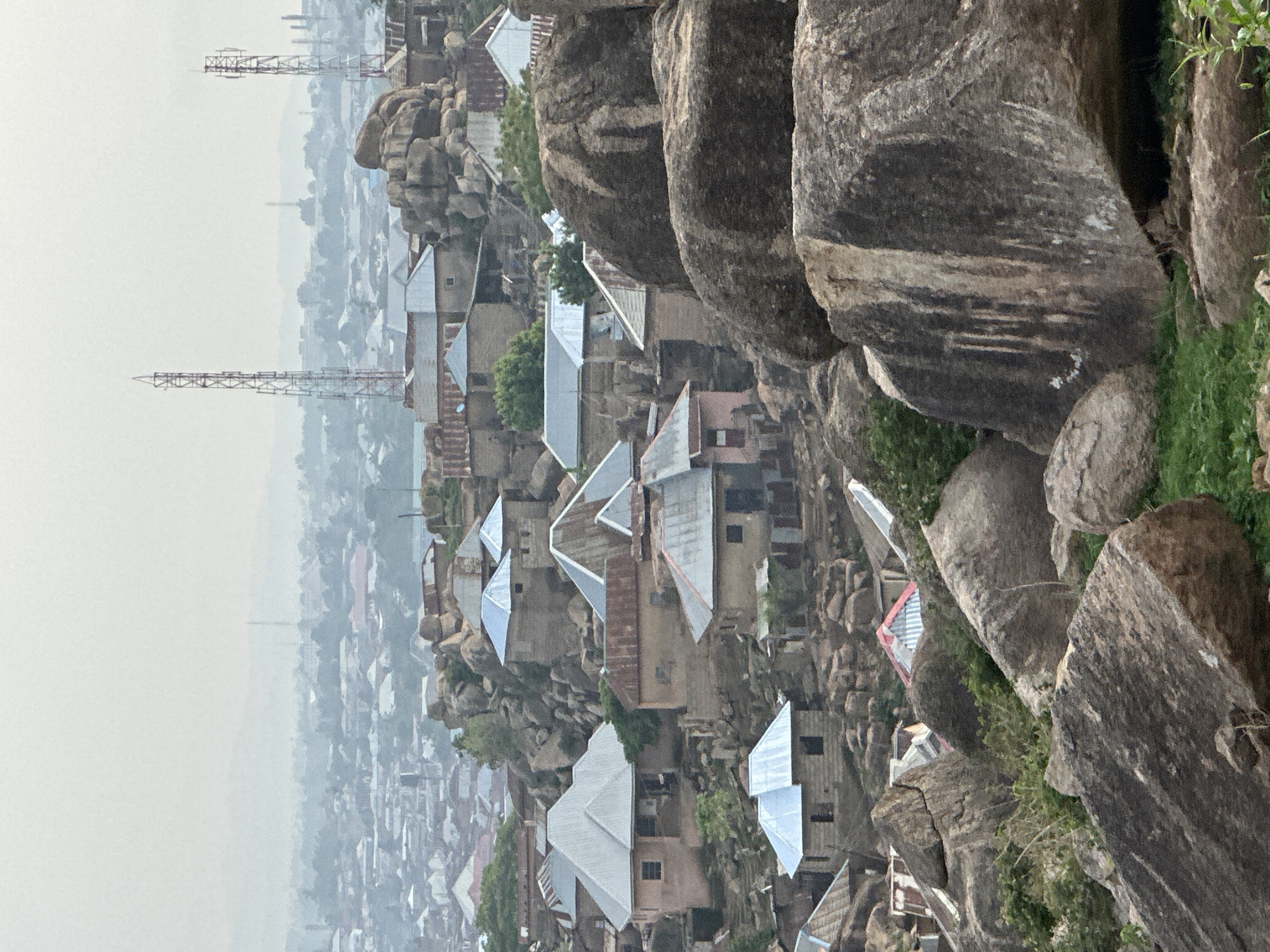 The image presents a panoramic view of a dense settlement situated on and around prominent, dark grey-brown rock formations, characteristic of the Jos Plateau in Nigeria. The perspective is from an elevated vantage point, looking down across the sprawling community.
In the foreground and midground, numerous houses are clustered between and atop the massive boulders. These structures primarily feature light-colored walls, likely concrete or mud brick, topped with various types of roofs. Many roofs are made of corrugated metal, some appearing new and shiny silver, while others are weathered and dull, showing signs of rust. Green foliage, including bushes and small trees, grows sporadically between the rocks and buildings, adding touches of nature to the rugged landscape. The houses appear to be built directly into the natural contours of the rocky terrain, showcasing adaptation to the environment.
Towards the background, the settlement extends into a hazy urban sprawl, with a muted cityscape fading into the atmospheric distance. Two tall, skeletal communication towers, resembling cell phone masts, stand prominently against the pale, overcast sky, indicating modern infrastructure within the developing area. The overall weather appears hazy or overcast, contributing to a soft, diffused light and reduced visibility in the furthest parts of the scene. No specific activities or people are discernible, and there is no visible text. The scene captures a moment of everyday life in a resilient community deeply integrated with its unique geological setting.