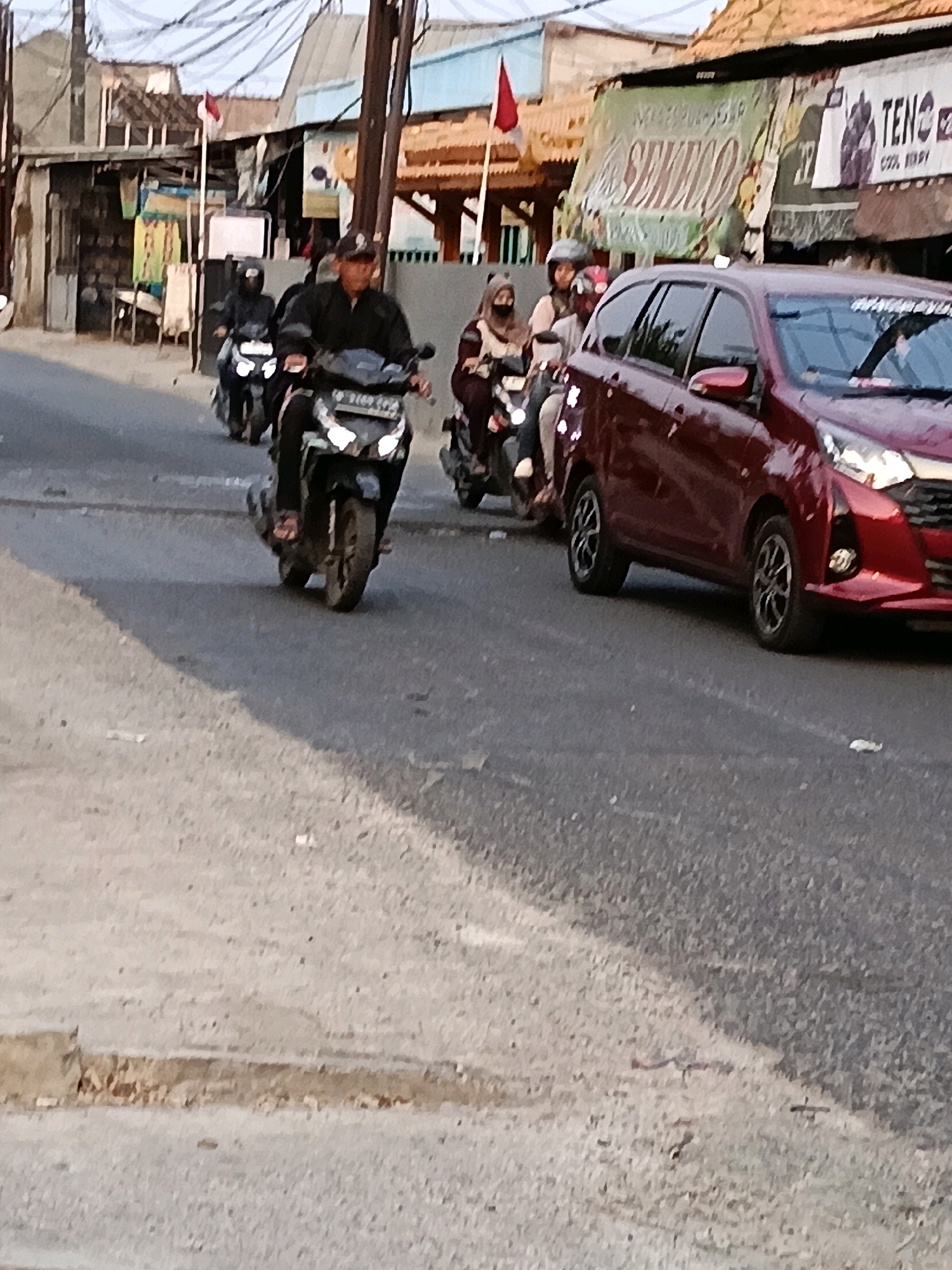 The image depicts a street scene in Belimbing, Indonesia. Several people are riding motorcycles in the foreground and mid-ground. A maroon car is parked or moving slowly on the right side of the road. Buildings with various signs and awnings line the street on the left. A red and white flag is visible on a pole in the background. The road surface appears to be asphalt. The lighting suggests it is daytime, possibly late afternoon due to the angle of the shadows.