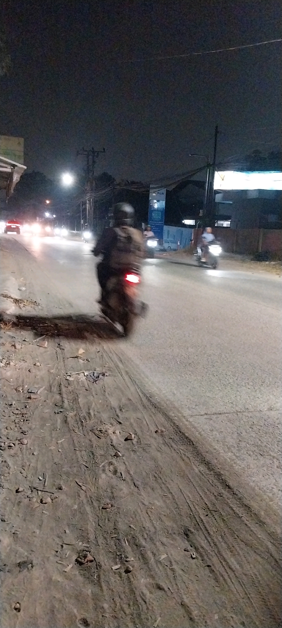 This is a nighttime photograph taken on a street in Kelapa Dua, Indonesia. The scene depicts a road with traffic, illuminated by streetlights and vehicle headlights. In the foreground, a dirt and debris-strewn shoulder is visible, with tire tracks imprinted on it. The main subject is a person riding a motorcycle, captured in motion with a blurred effect, showing the red taillight. Further down the road, other vehicles, including motorcycles and cars, are also visible, their headlights creating streaks of light. Several utility poles with wires are present along the roadside, and a sign with the "PLN" logo (Perusahaan Listrik Negara, the Indonesian state electricity company) is visible in the background. The environment appears to be a moderately developed urban or suburban area, with buildings and trees lining the street. The overall impression is a typical street scene at night in Indonesia.