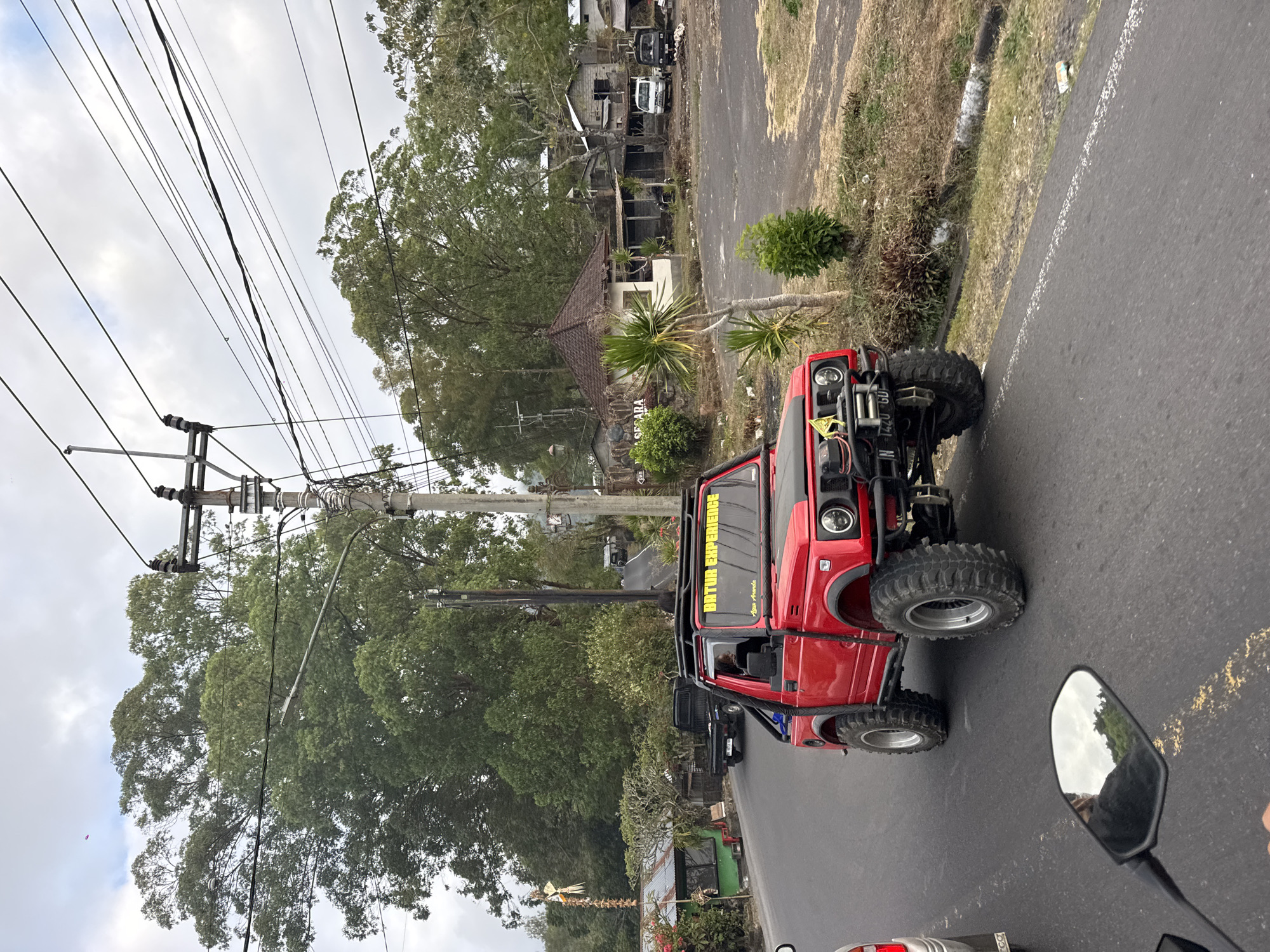 In Kintamani, Indonesia, a bright red, off-road Suzuki Samurai is traveling on a paved road under an overcast sky. The vehicle is heavily modified with large tires, a roof rack, and a winch. The driver is partially visible inside. In the background, several buildings and trees line the side of the road, and a utility pole with numerous wires stands prominently. Further in the distance, more trees and a few parked vehicles are visible. The perspective suggests the photo was taken from another moving vehicle, possibly a motorcycle, as a portion of a rearview mirror is in the foreground.