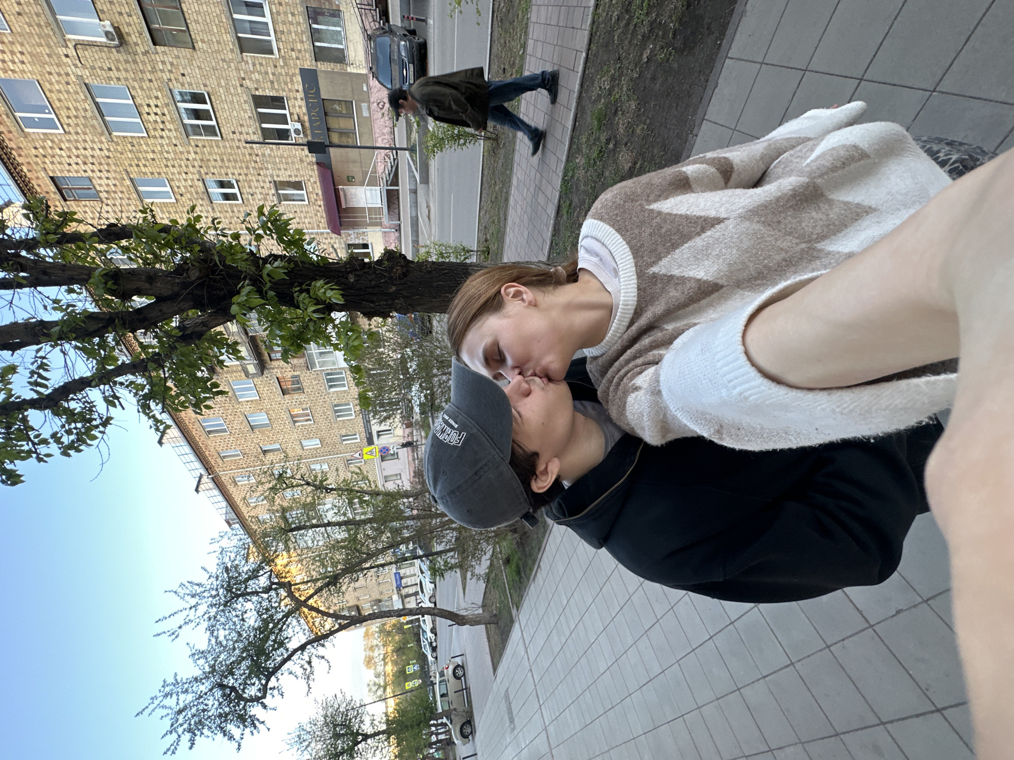 A photo taken at an angle from above captures two people kissing on a sidewalk beside a tree-lined street. One person is wearing a light-colored sweater with a diamond pattern and the other is wearing a dark-colored hoodie and baseball cap. The background features a multi-story brick building and a pedestrian crossing the street. The location appears to be Krasnoyarsk, Russia, based on identifiable landmarks in the surrounding environment.