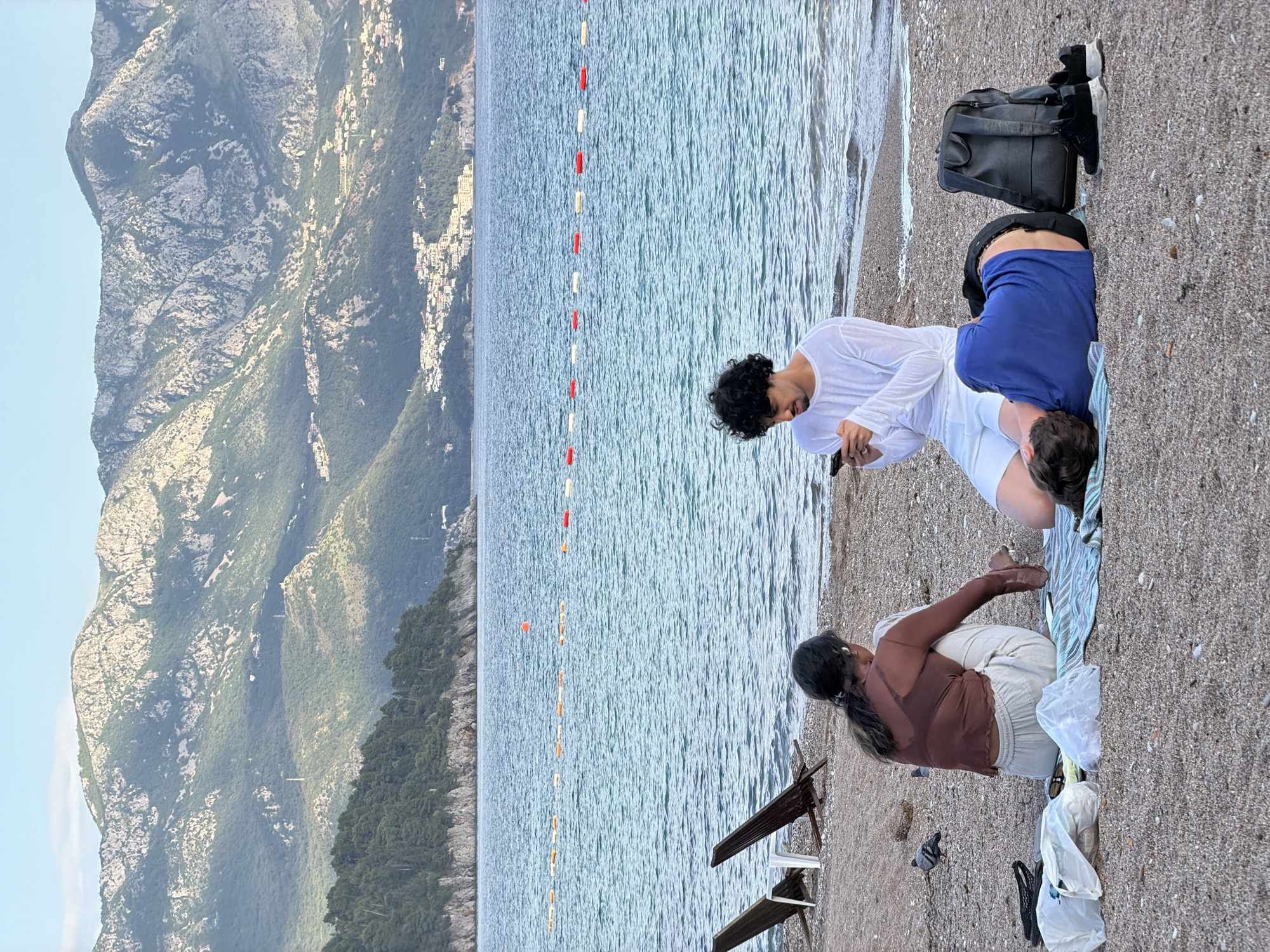 The image depicts a scene on a beach near Budva, Montenegro. In the background, there is a large mountain range. The foreground shows several people on a sandy beach. A series of red and white buoys are visible in the water. One person in a white shirt is standing and looking at a phone. Another person in a blue shirt is reclining on a beach towel. A third person is sitting on the sand. There is a black backpack and other personal items nearby. There are some chairs on the beach, closer to the camera.