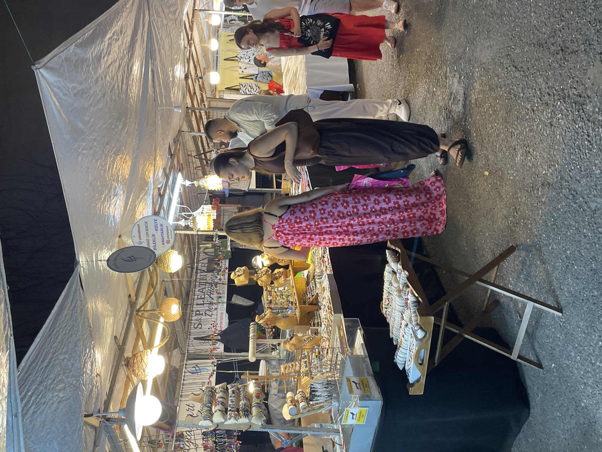 This media file captures a lively outdoor market scene, likely at dusk or night, given the artificial lighting. The primary subjects are several people browsing and interacting at a vendor's stall. A woman in a red dress stands at the back, while a couple in the foreground is looking at items displayed on a table. Another woman in a pink floral dress is also engrossed in examining the merchandise.
The stall itself is laden with various handcrafted items, including an abundance of colorful bracelets, necklaces, and possibly other accessories. Some items are displayed on mannequins, and there are also smaller displays of jewelry and decorative objects. The vendor's sign, partially visible, features text including "S&P LEATH," "yukselglassart," and "MURANO CAM BONCUK BİLEKLİK KOLYE ANAHTARLIK," suggesting the sale of leather goods, Murano glass beads, bracelets, necklaces, and keychains. The setting appears to be a street or market area, with a textured asphalt ground. The atmosphere seems relaxed and engaging, with people enjoying their time at the market. The presence of overhead lighting and a large white tarp suggests an evening event or a covered market area.