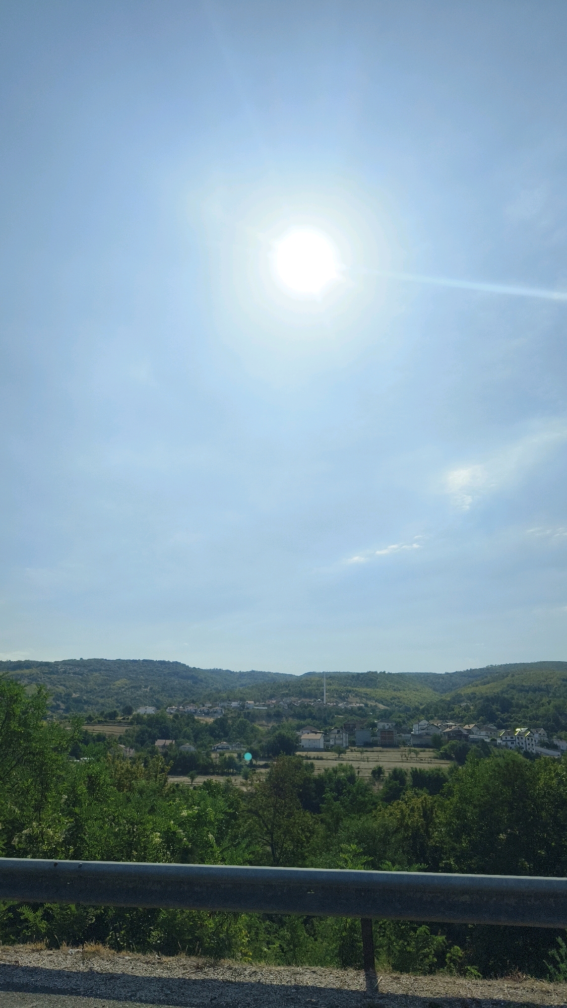 The image captures a bright, sunny day overlooking a valley with a village. The sun is high in a clear blue sky, with a slight haze. In the foreground, a metal guardrail runs horizontally across the lower portion of the image, with dry grass and gravel on the ground beneath it. Behind the guardrail, lush green trees and foliage dominate the mid-ground, forming a dense forest. Further back, nestled within the valley, lies the village of Novo Selo, North Macedonia. Several buildings, primarily houses with white and light-colored walls and tiled roofs, are visible, interspersed with more greenery. A tall, slender industrial chimney or tower stands out amongst the village buildings. The landscape is characterized by rolling hills covered in trees that extend into the background, creating a sense of depth. The overall scene is peaceful and rural.