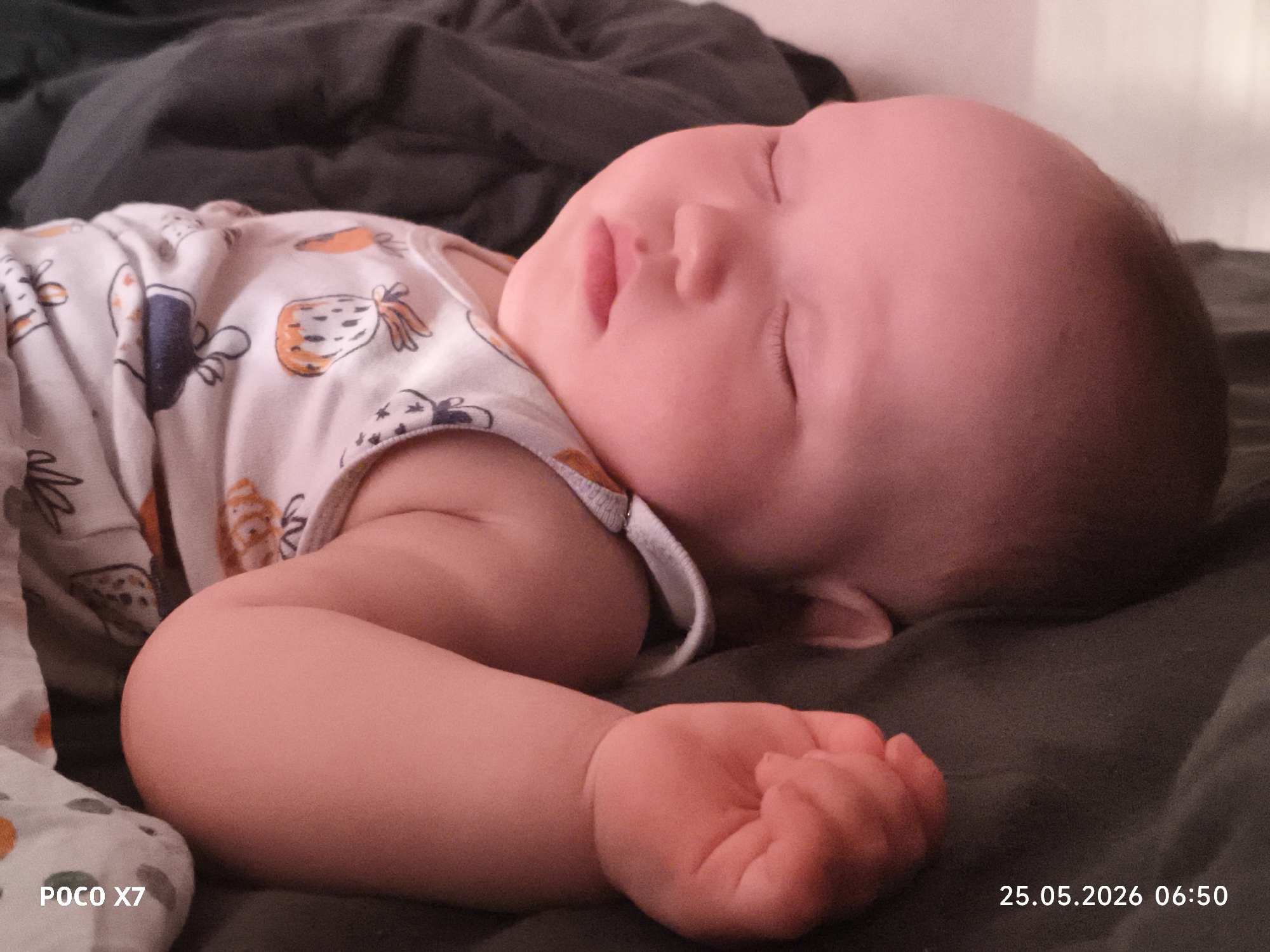 A baby is sleeping on a dark-colored surface. The baby is wearing a light-colored patterned shirt with various designs. The baby's eyes are closed, and its mouth is slightly open. The baby's left arm is bent with its hand near its face. The background is slightly blurred. The location and city are unknown. The date and time "25.05.2026 06:50" and "POCO X7" are visible at the bottom of the image.