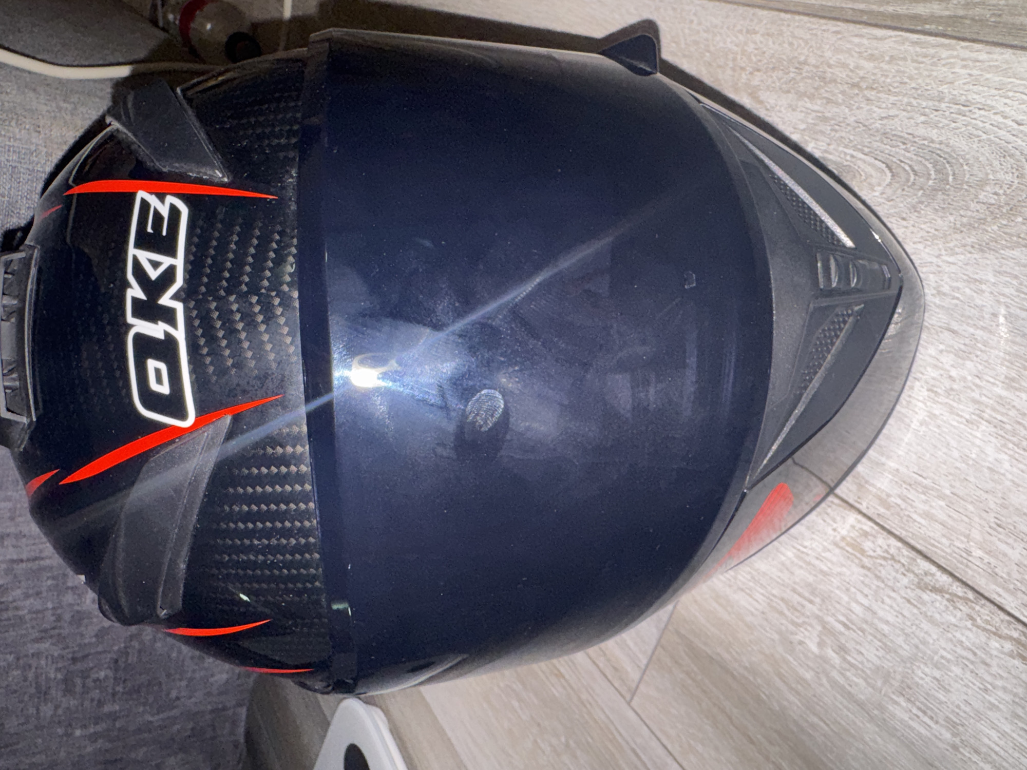 A close-up, eye-level, frontal shot shows a black and red motorcycle helmet with a dark visor, resting on a wooden floor. The helmet features carbon fiber-like detailing on the back and a white "OKE" logo on the side. Red racing stripes adorn the top and sides. The background is blurred, showing a textured grey surface and a hint of what appears to be a grey sofa. The lighting suggests an indoor setting, likely a room in Bishkek City, Kyrgyzstan, possibly a home or storage area. There are no people visible in the image. The overall impression is one of an object placed for display or storage.