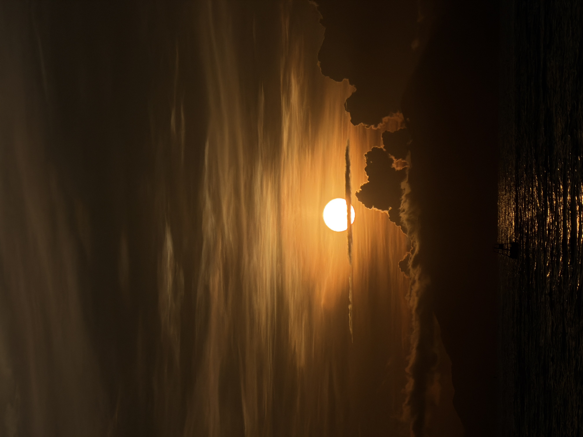 The image depicts a sunset over the ocean near Hoi An, Vietnam. The sun, a bright orb of light, is partially obscured by clouds, casting a warm, golden hue across the sky. The clouds are a mix of dark, dramatic formations and wispy, translucent layers. In the distance, the silhouette of a mountainous coastline is visible. The ocean surface is dark and textured, reflecting the sunset's glow with shimmering highlights. A small sailboat with a single sail is positioned in the lower right quadrant of the image, appearing as a dark silhouette against the water's reflection. The overall scene conveys a sense of tranquility and natural beauty.
