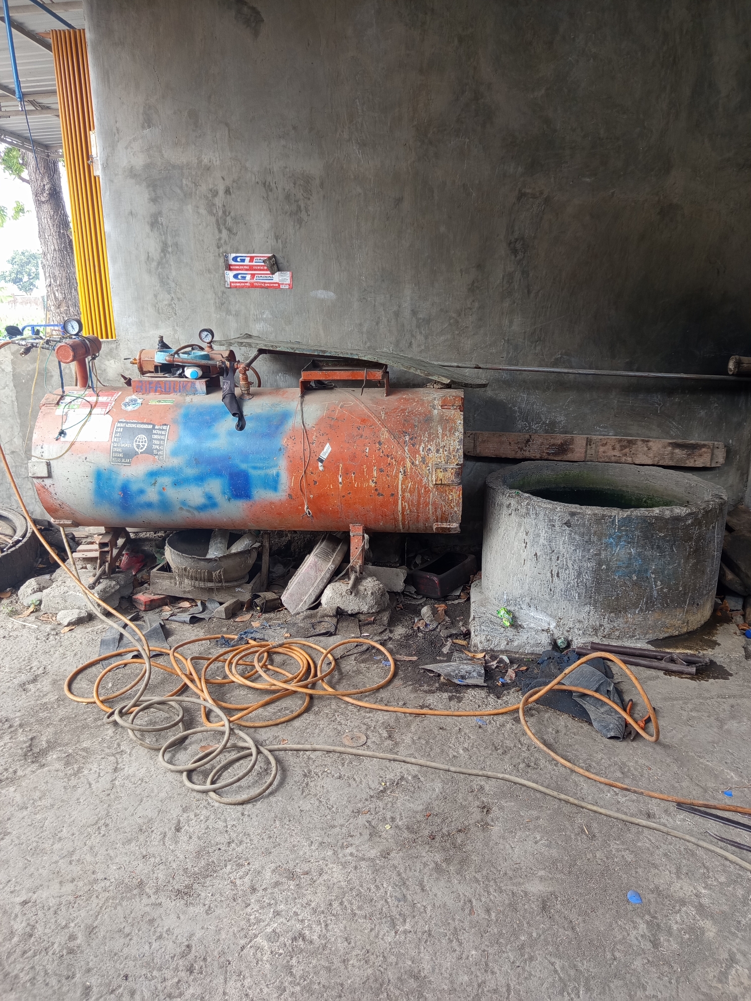 In Mrican, Indonesia, an outdoor workshop scene unfolds. A large, aged, orange and grey cylindrical air compressor dominates the center, showing signs of rust and wear, with a blue painted section on its side. It rests on makeshift supports, surrounded by various tools and debris, including coiled hoses, a concrete well with murky water, and scattered bricks and metal scraps. The workshop is situated against a rough concrete wall, with a yellow accordion-style door visible on the left, partially obscuring a tree. Two small rectangular stickers with red and white lettering are affixed to the concrete wall above the air compressor. The ground is a rough, grey concrete surface, littered with more debris. The overall impression is one of a functional, albeit cluttered and weathered, automotive or industrial repair space.