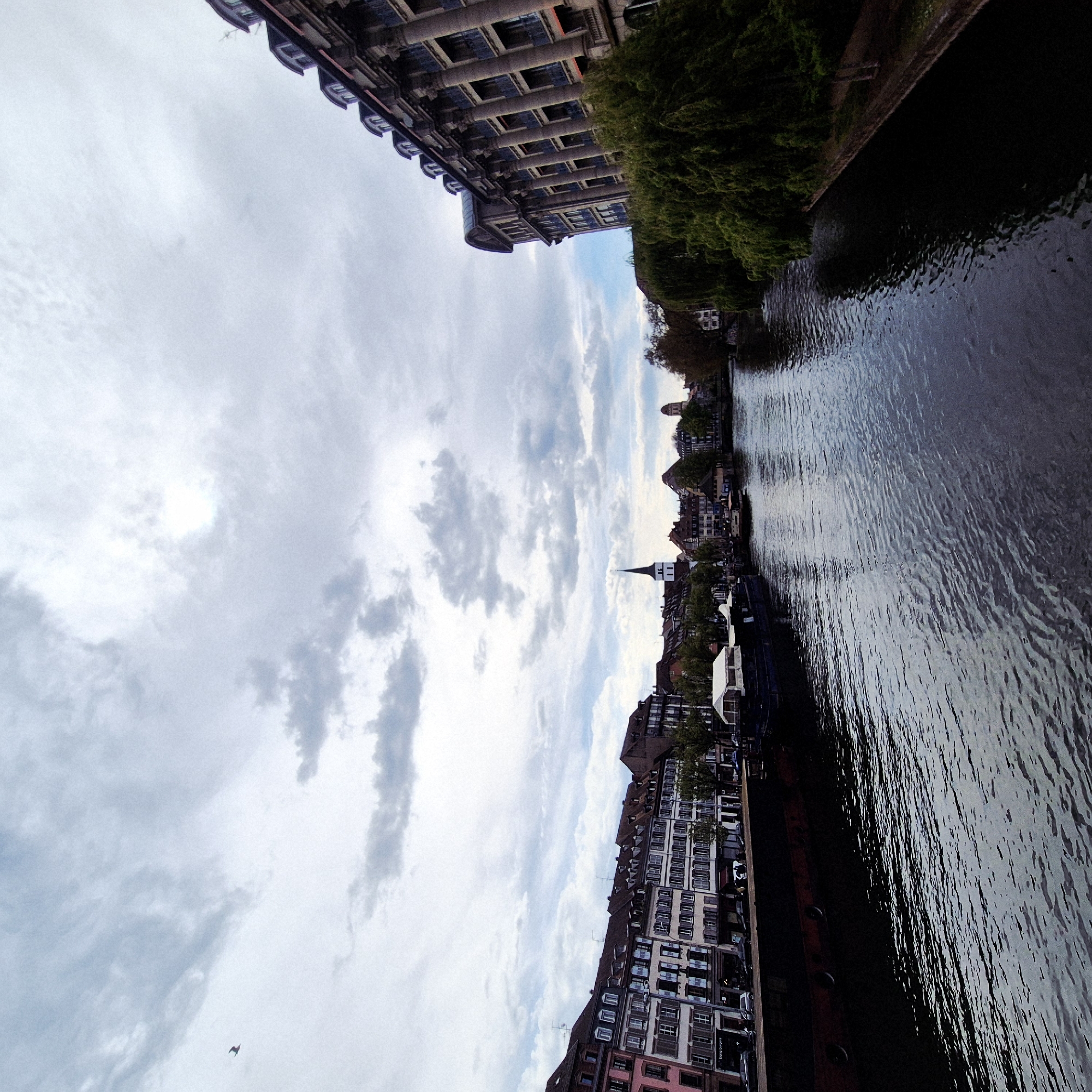 The image presents a view in Strasbourg, France, beside a canal. The right side shows a canal with water. On the left side, there are buildings including a spire. Above are some clouds and sky.