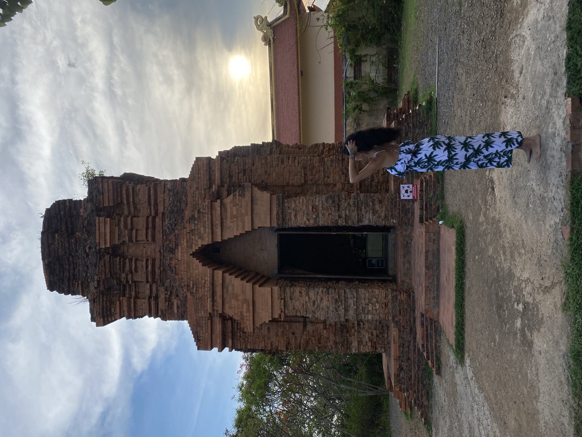 The image shows an ancient brick tower located in Phu Thuy Ward, Phan Thiết, Bình Thuận, Vietnam, standing beside a temple. In the foreground, a woman with long dark hair and a dress decorated with palm trees poses in front of the tower. The tower is made of dark red bricks, with a series of staggered ledges and a doorway leading into a dark interior. The sky is visible behind the tower, with the sun creating a bright glare. A gravel path runs alongside the tower, bordered by grass.