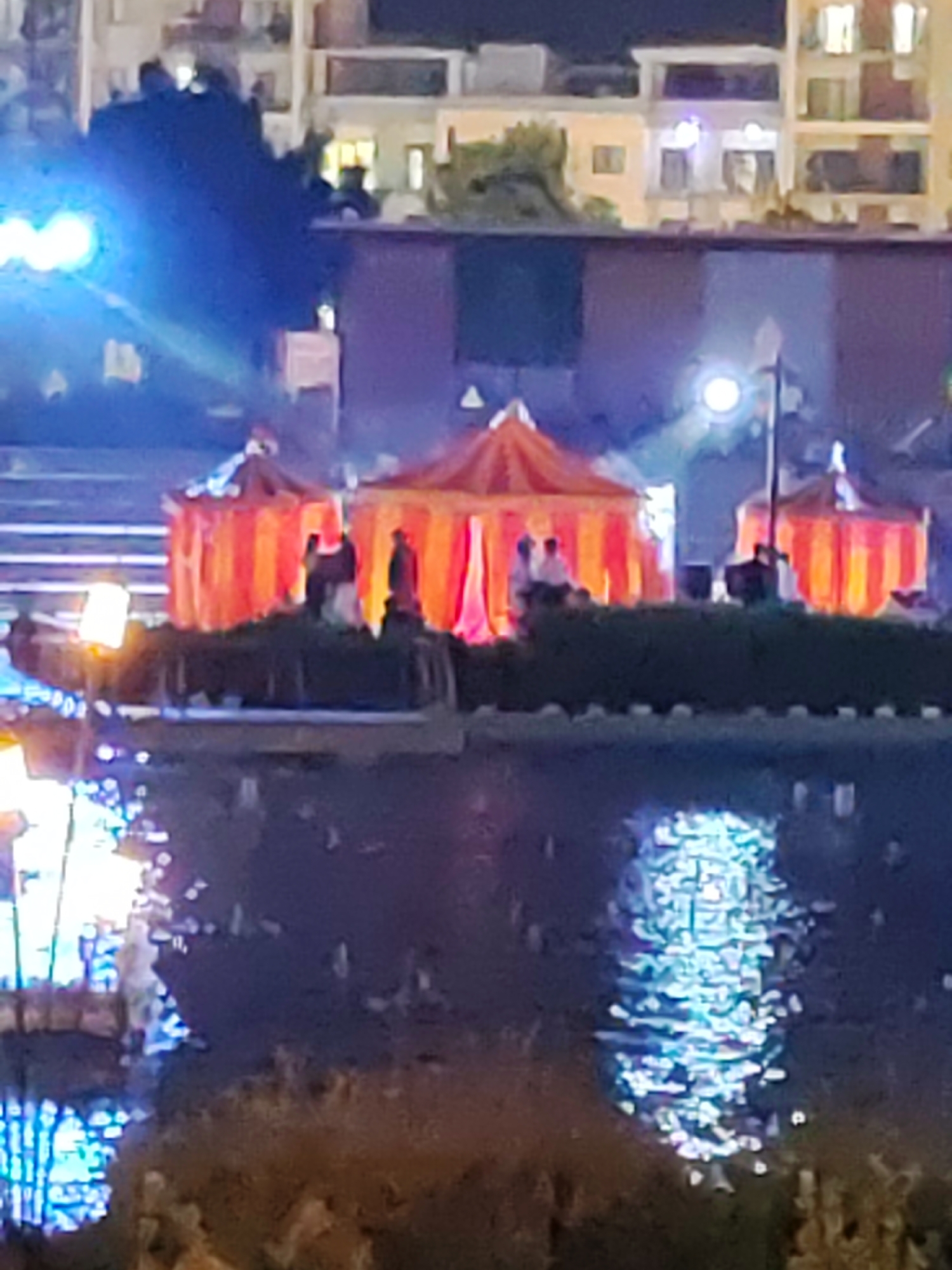 The image shows a night scene with several illuminated tents, likely part of a festival or event, positioned along a waterfront. The tents are orange and yellow striped and appear to be temporary structures. There are people visible within and around the tents, some standing and some sitting. The waterfront is reflecting the lights from the tents and the surrounding buildings, creating a shimmering effect on the water. In the background, there are multi-story buildings with illuminated windows. The overall atmosphere suggests a lively evening gathering.
This scene is consistent with events held at the Tashkent Botanical Garden, which features a lake and hosts various festivals and light installations, particularly during evening hours.