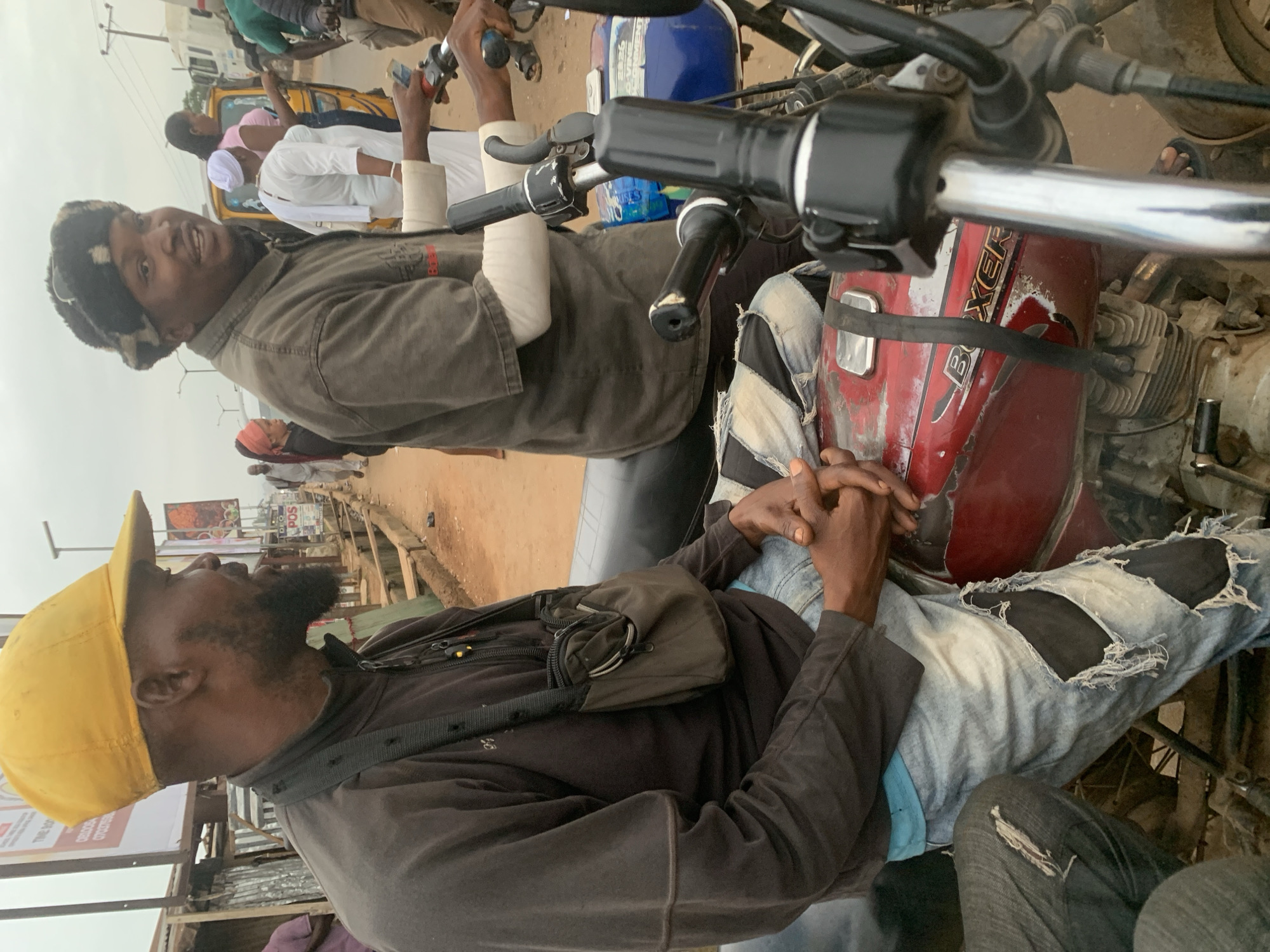 The image depicts an outdoor scene in the city of Shagamu, Nigeria. In the foreground, two men are seated on a red motorcycle. The man on the left is wearing a yellow baseball cap and a dark long-sleeved shirt, with a small brown bag hanging around his neck. He is also wearing distressed light-blue jeans with large rips. His hands are clasped in his lap. The man on the right is wearing a dark, possibly olive-colored, long-sleeved shirt and a dark, fur-lined hat. He has a white band around his right forearm and is holding the motorcycle's handlebars. The motorcycle has a red fuel tank with the word "BOXER" visible. In the background, other people and vehicles are present, including a yellow tricycle and a white truck. The environment appears to be a street with a dusty, reddish-brown ground. There are several signs and structures visible along the side of the road, suggesting a commercial or market area. The sky is overcast.