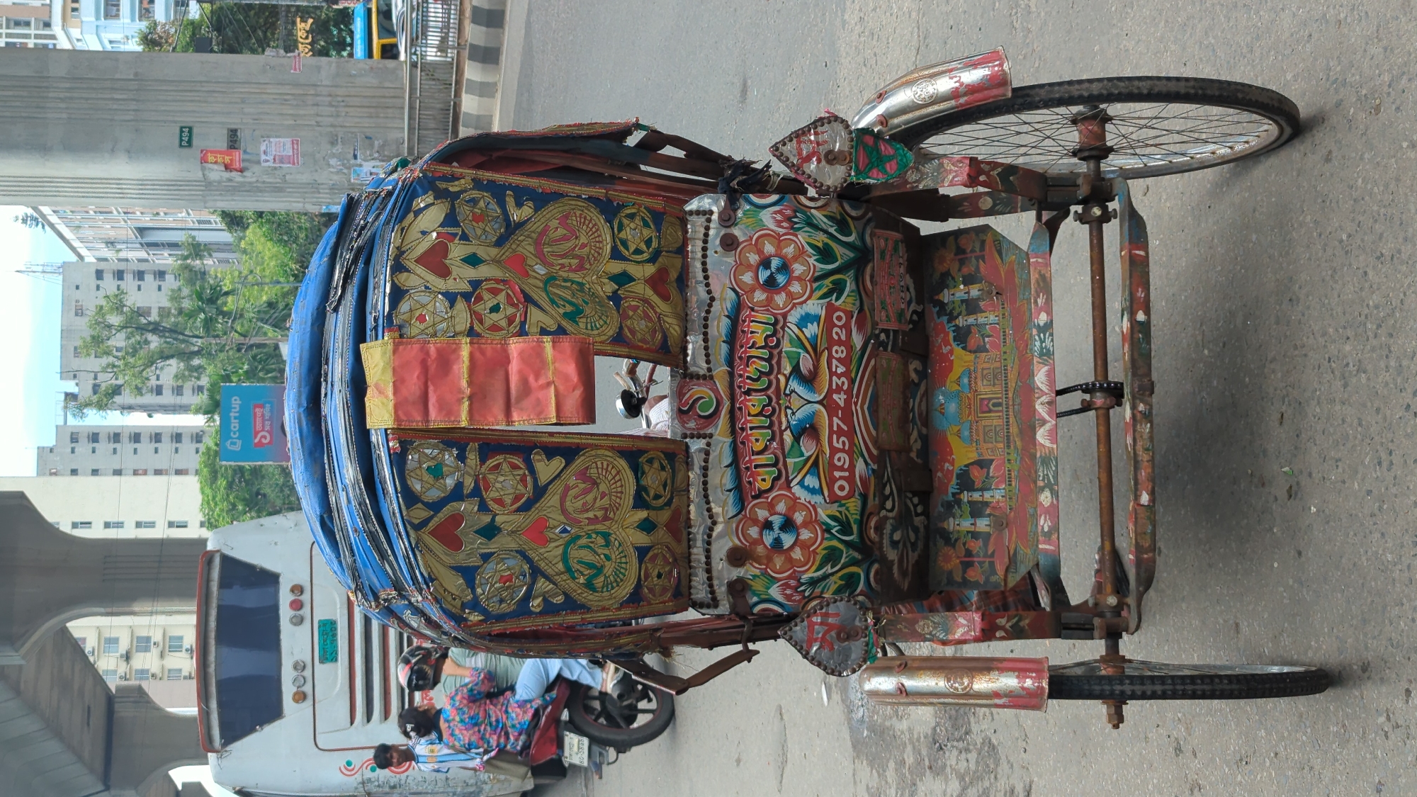 This is a forward-facing, medium shot of a brightly decorated auto-rickshaw in Dhaka, Bangladesh. The auto-rickshaw is parked on a paved road with a pedestrian walkway on the right side. The auto-rickshaw has intricate designs painted on its rear and sides, featuring floral patterns, birds, and what appears to be a depiction of a mosque. A phone number, "01957-437820," is clearly visible on the side. In the background, a white bus is seen, along with several tall buildings and an elevated roadway. A person on a motorcycle is visible to the left of the auto-rickshaw. The sky is overcast.