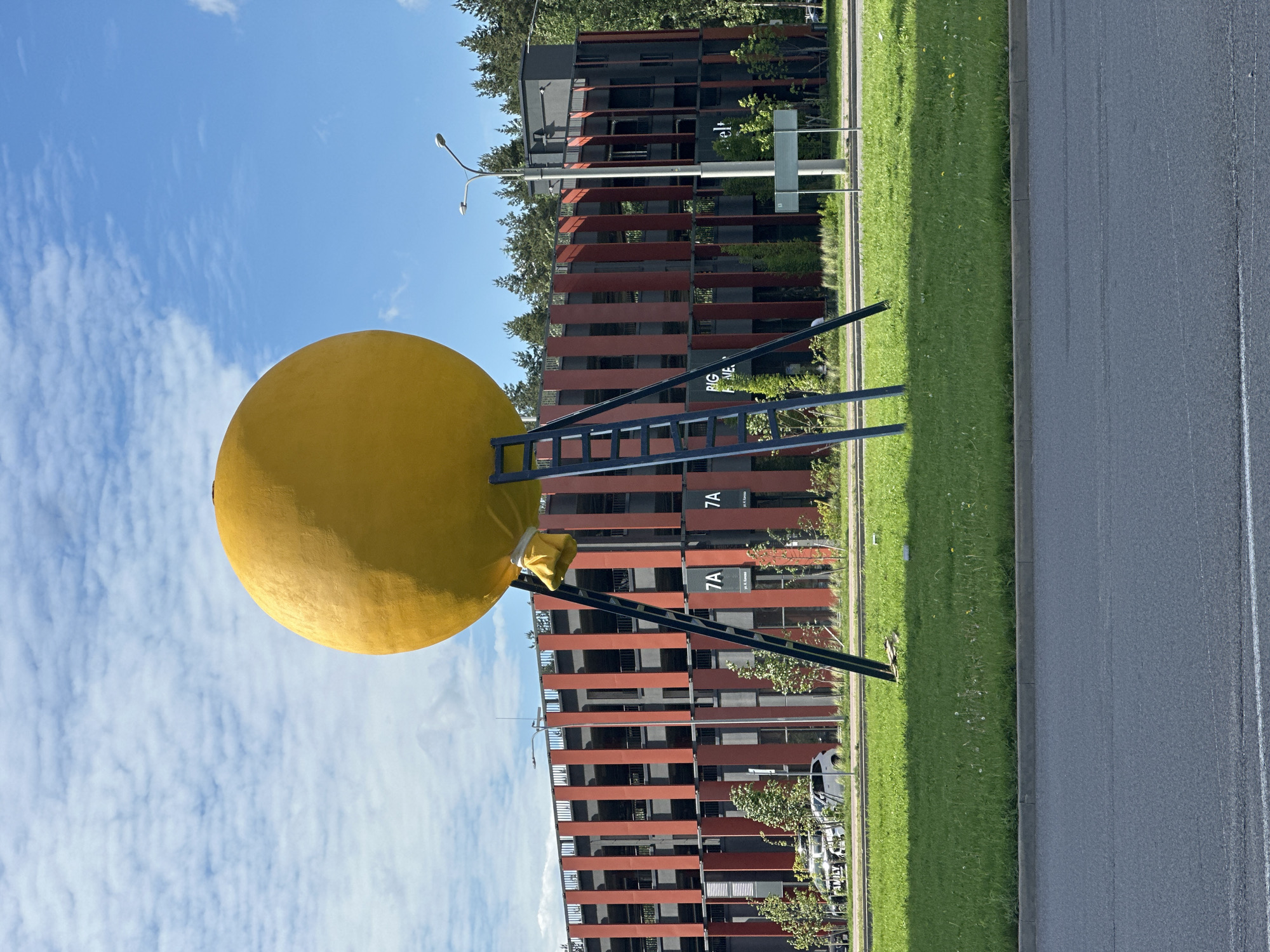 A yellow spherical sculpture rests on a metal frame, resembling a ladder, located in Бараўлянскі сельскі Савет, Belarus. Behind the sculpture stands a modern apartment building with dark, horizontally-oriented panels on its facade. The sculpture is set on a grassy lawn next to a paved road. The sky above is light blue with scattered white clouds. A street lamp and street signs are visible in the background.