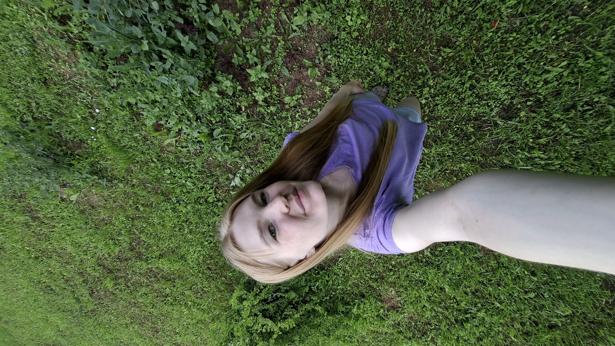 A young woman with blonde hair is taking a selfie outdoors. She is wearing a purple t-shirt and light blue shorts. The woman is standing on a grassy area. The background is filled with green grass and some bushes. The lighting suggests it is daytime. The location is identified as Bolshoy Bukor, Russia.