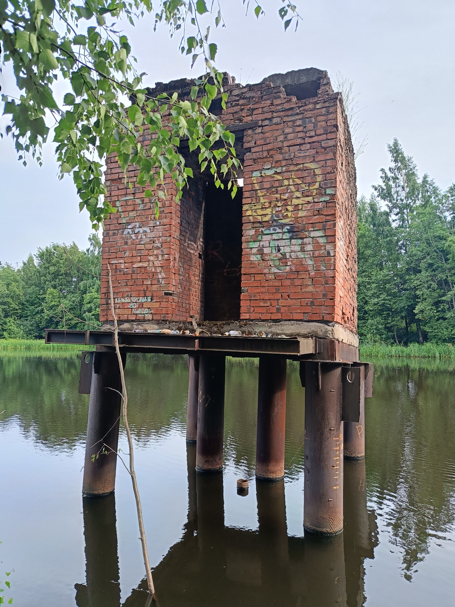 The image depicts a derelict brick structure standing on stilts over a body of water, surrounded by trees. The structure appears to be a small building, possibly a pump house or control station, with an open doorway and visible graffiti on its exterior. The brickwork is weathered and some of the top is missing, suggesting it is abandoned. It is supported by thick, rusty metal pillars that extend into the water. The water is calm, reflecting the overcast sky and the surrounding greenery. A thin, bare branch leans against the structure, adding to the sense of decay. In the background, lush green trees form a dense forest line. The overall mood is one of abandonment and nature reclaiming a man-made structure. The time of day appears to be overcast, possibly morning or late afternoon, given the diffused light. The location is identified as Saint Petersburg, Russia. There is some graffiti on the brick walls, but it is not clearly legible.