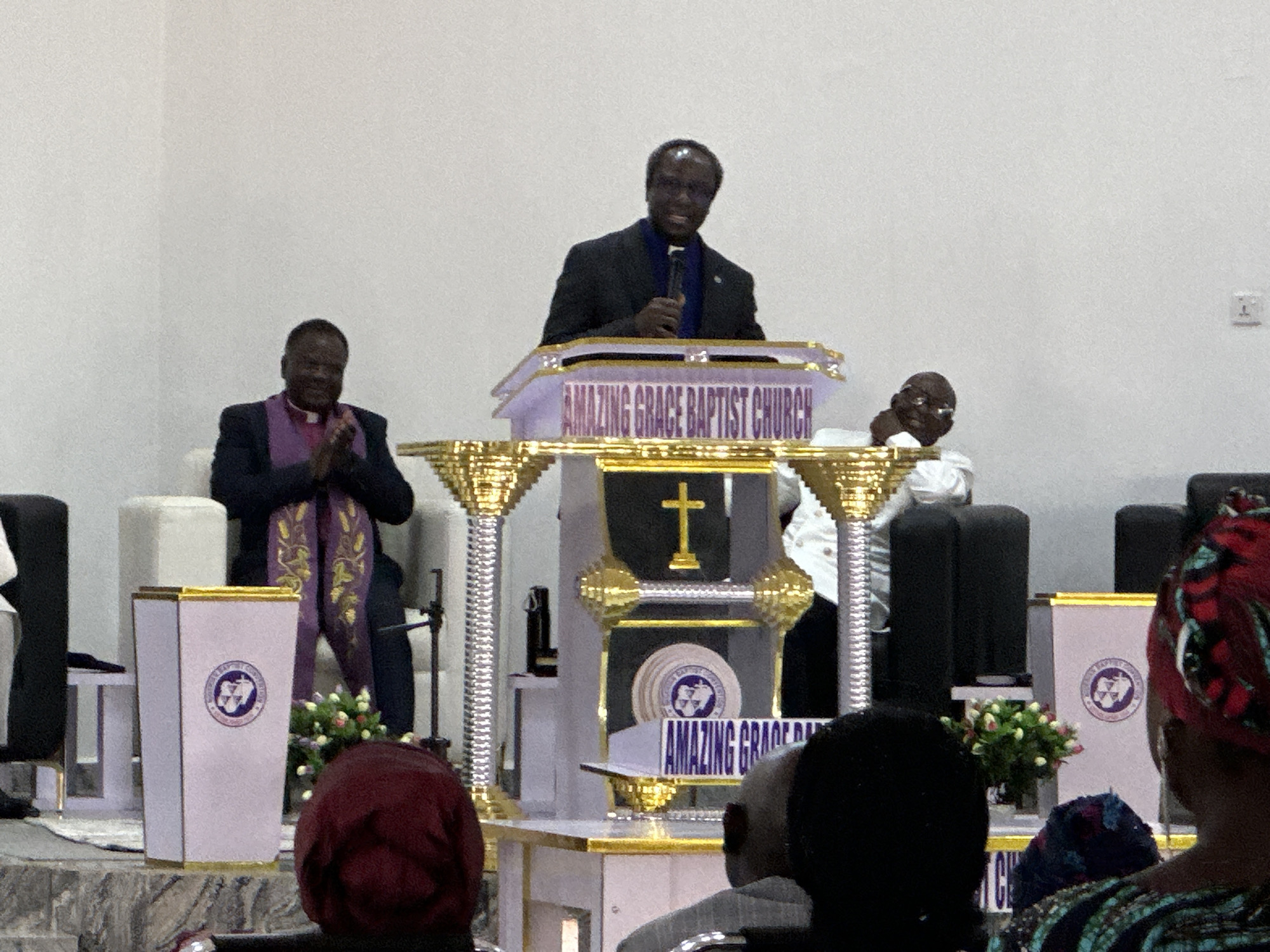 The image displays an indoor scene at the Amazing Grace Baptist Church in Goni Gara, Nigeria. A dark-skinned male subject, wearing a dark suit and a blue shirt, stands centrally at an ornate white and gold podium, holding a black microphone in his right hand. The podium features "AMAZING GRACE BAPTIST CHURCH" inscribed in purple text across its front.
To the left of the speaker, a dark-skinned male subject is seated, wearing a dark suit with a purple and gold patterned stole draped over his shoulders, his hands clasped together. To the speaker's right, a dark-skinned male subject with a bald head, dressed in a white shirt, is partially visible, also seated.
The stage area includes a decorative structure behind the speaker, characterized by gold pillars, a prominent golden cross, and circular elements displaying the church's logo. Two identical white and gold pedestals, each adorned with the church's logo and a small arrangement of white and red flowers, are situated on the stage. White upholstered chairs are visible behind the seated individuals. The background is a plain white wall, with a single electrical outlet positioned on the right side. In the immediate foreground, the backs of several audience members' heads are visible, some wearing patterned head coverings.