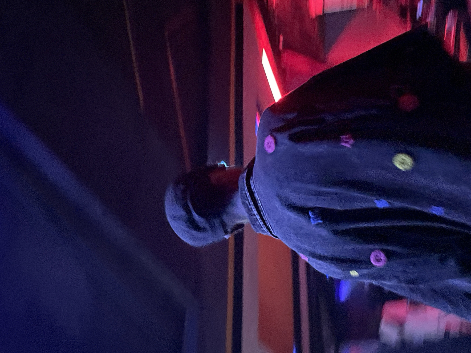 The image shows a person from behind, wearing a dark jacket decorated with colorful circular and square patches. The person is also wearing a dark cap. They are standing in a dimly lit environment with strong red and blue lighting. The background appears to be indoors, with horizontal bands of orange and yellow visible on a wall. The scene is consistent with being in a club or a venue with dynamic lighting. The city of Durumi, Nigeria is not depicted in this image.