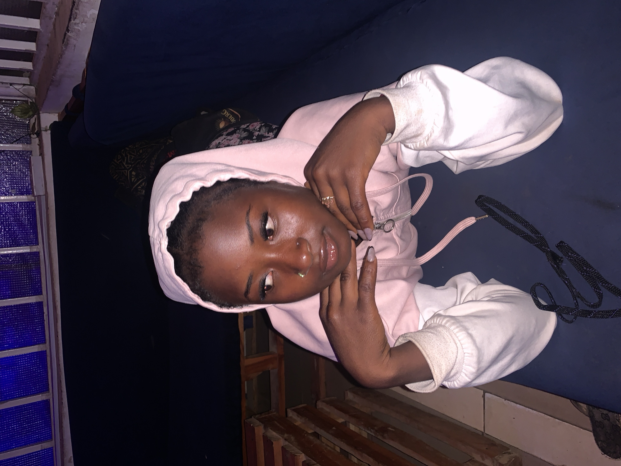 An intimate, slightly angled portrait captures a young individual, likely female, with dark skin, wearing a light pink hooded sweatshirt. She is resting or leaning back on a dark blue surface, possibly a sofa or bed. Her short, dark hair is visible beneath the hood. She looks towards the camera with a subtle, composed expression, highlighted by a nose piercing and glossy lips. A ring adorns her right hand, and her nails are painted a light, neutral shade.
The scene is dimly lit, suggesting it is evening or night. To the left of the frame, architectural elements featuring blue, textured panels and a small green plant are visible. Below the subject, a series of wooden slats or planks indicate a wooden structure or flooring. Behind her head, a dark, patterned fabric object is partially seen, on which the text "BERG" is discernable. The individual's hands are casually positioned near her chest, with pink and darker drawstrings from her hoodie visible. The overall ambiance is relaxed and casual. This image is situated in Barakin Akawo, Nigeria.