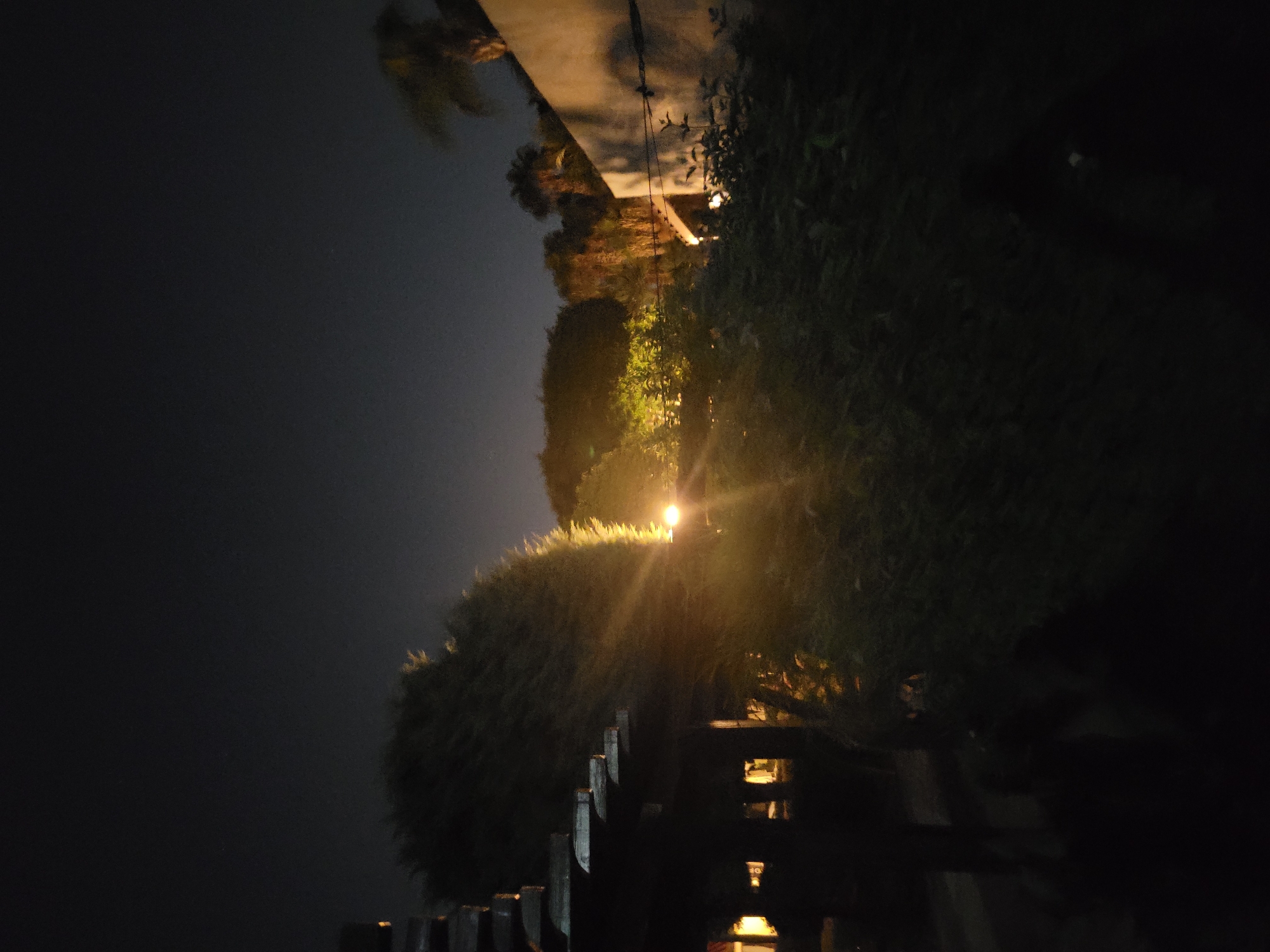 This is a nighttime outdoor photograph taken in Trachoni, Cyprus. The image depicts a dimly lit garden or courtyard area. In the foreground, a dark, wooden structure, possibly a pergola or awning, is visible on the left. Beyond this structure, dense green foliage fills the mid-ground, with a bright, yellowish light source creating a lens flare effect that beams across the vegetation. Further in the background, trees and palm trees are silhouetted against a dark night sky. To the right, a light-colored wall with shadows cast upon it is visible, suggesting an adjacent building. The overall atmosphere is serene and quiet, with artificial lighting illuminating parts of the scene.