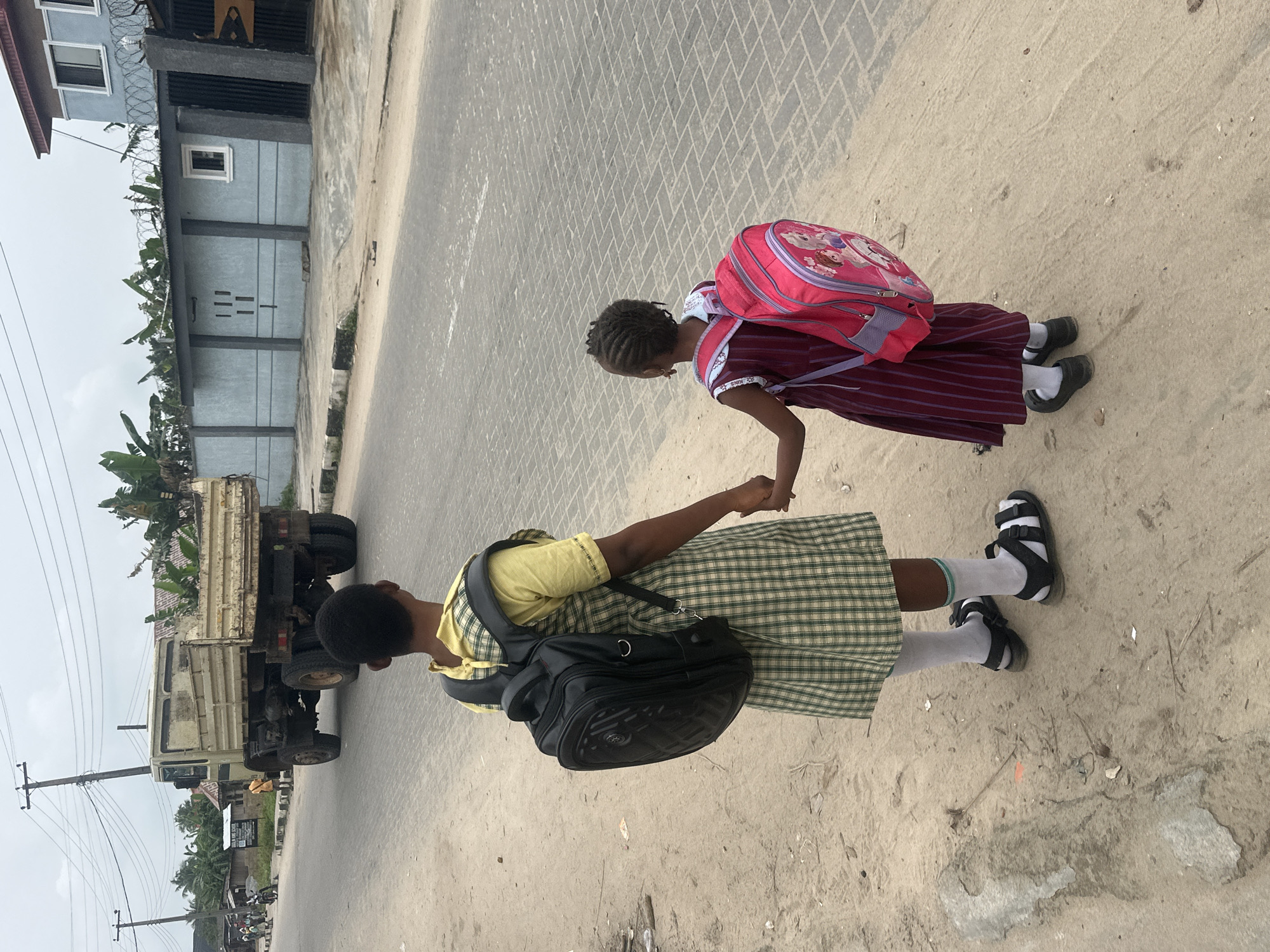 In Badagry, Nigeria, two young girls, likely students, stand on a dirt roadside holding hands. The older girl on the left wears a green and yellow plaid school uniform, white socks, and black sandals, with a large black backpack. The younger girl on the right wears a maroon striped dress, white socks, and black sandals, carrying a bright pink backpack adorned with cartoon characters. Behind them, a beige dump truck is parked on the paved road. The environment includes a sidewalk, a grey concrete wall with a corrugated metal roof, and lush green vegetation. The sky is overcast.