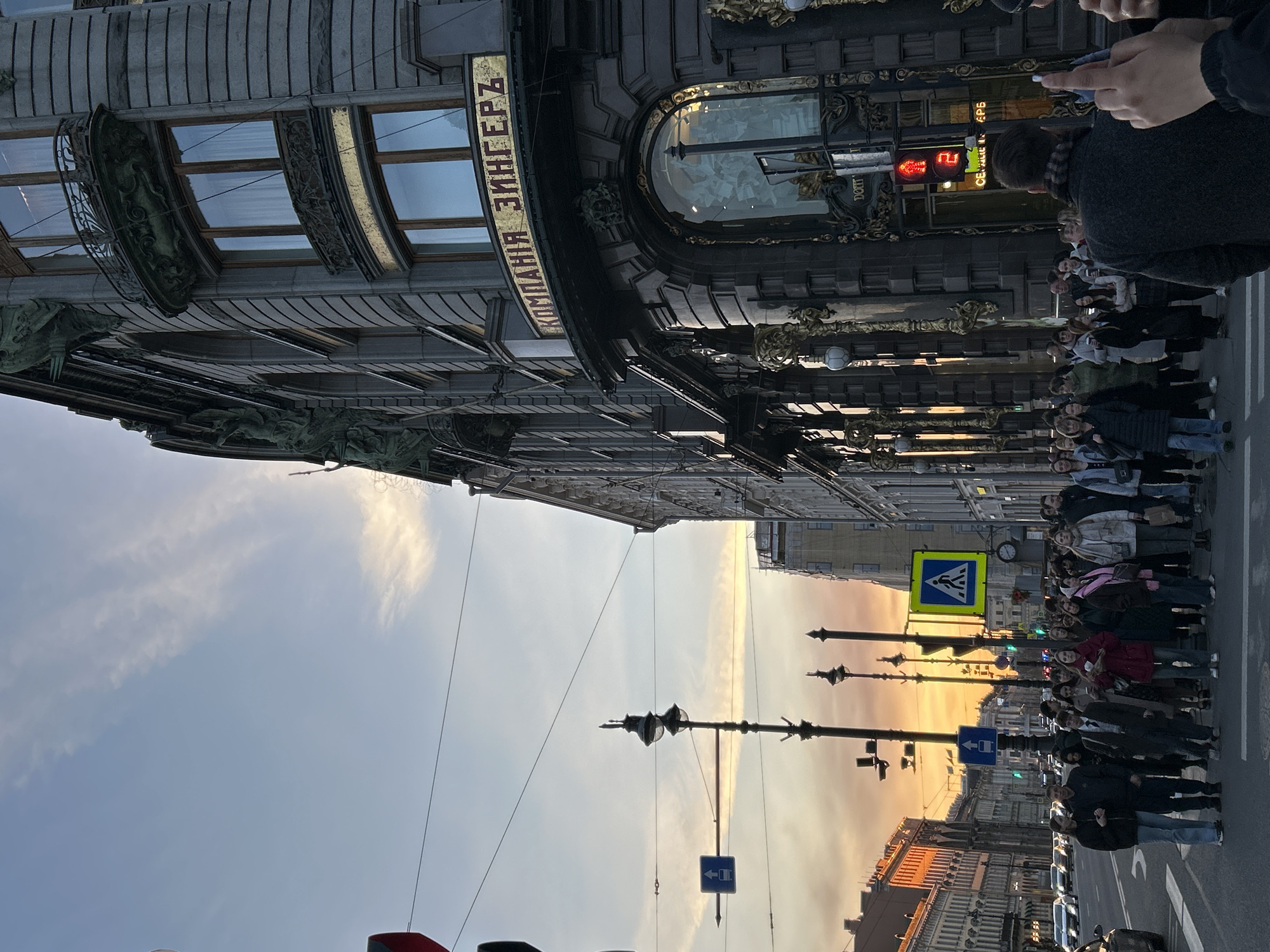 An urban street scene unfolds during what appears to be sunset or sunrise, casting a warm, golden light across the horizon and reflecting on buildings. The prominent subject is a grand, ornate building on the left, identified by the Cyrillic text "КОМПАНИЯ ЗИНГЕРЪ" (Singer Company) on its facade, strongly suggesting the Singer House (Dom Knigi) in Saint Petersburg, Russia. This building features large, elaborately framed windows, decorative metallic elements, and sculptural details.
On the right side of the street, a group of approximately 20-30 people, mostly adults, stand gathered on the sidewalk. They appear to be waiting to cross, as a pedestrian crossing sign (yellow and blue) and traffic lights are visible in the middle ground. One traffic light, closer to the camera, shows a red signal with a countdown number "2". Streetlights and various street signs, including a blue 'one-way' or 'keep left' arrow sign, line the street. Cars are also visible further down the road. The sky overhead transitions from a clear blue to vibrant hues of orange and yellow near the horizon, with some wispy clouds. The overall atmosphere is that of a bustling city transitioning from day to evening.