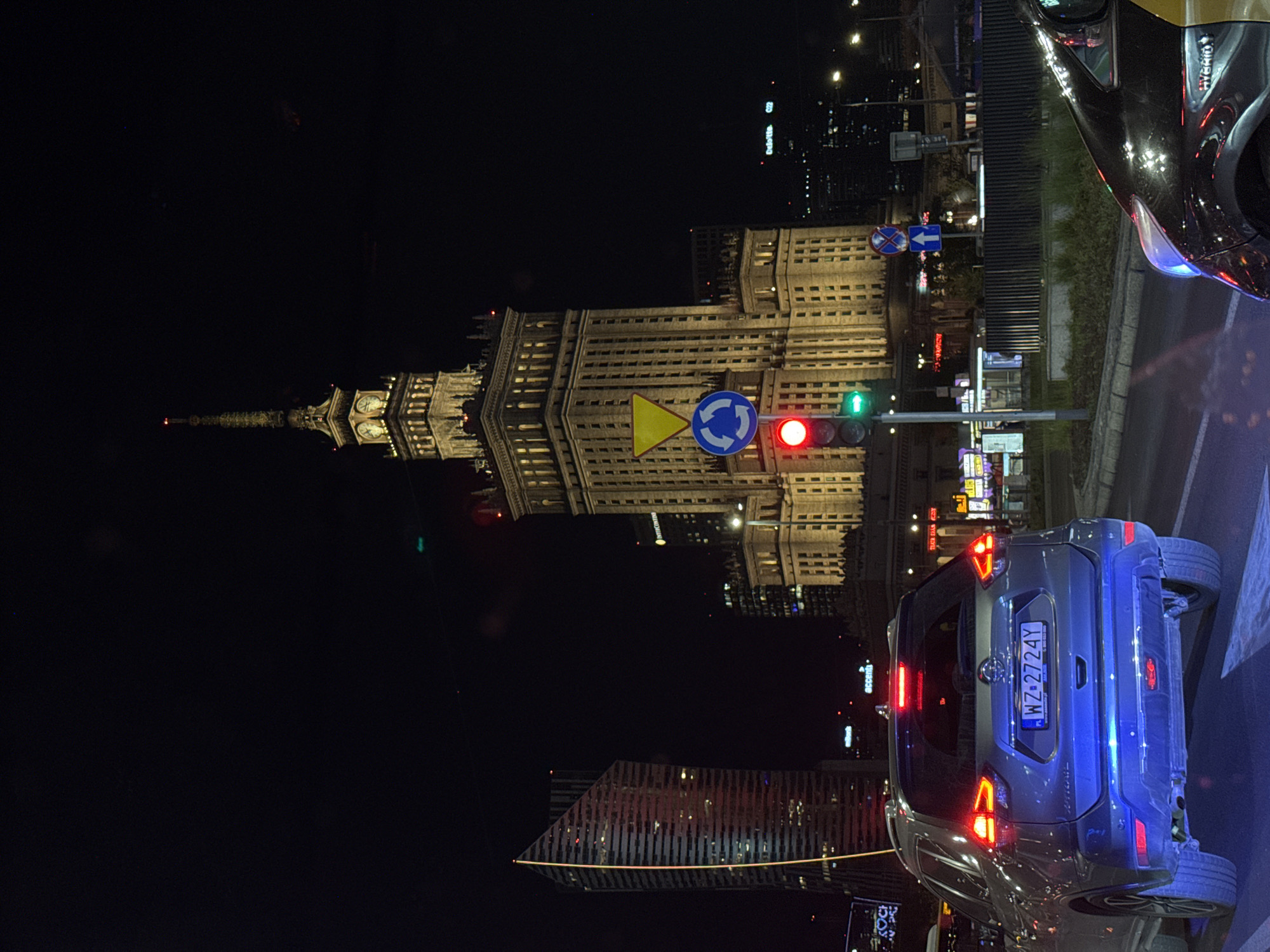 The image captures a nighttime scene in Warsaw, Poland, prominently featuring the Palace of Culture and Science illuminated in the background. In the foreground, a blue car with the license plate "WZ 2724Y" is visible, along with a portion of a black car. A traffic light shows red, and several street signs are present, including a roundabout sign and a yield sign. Modern buildings with illuminated windows contribute to the cityscape in the background.