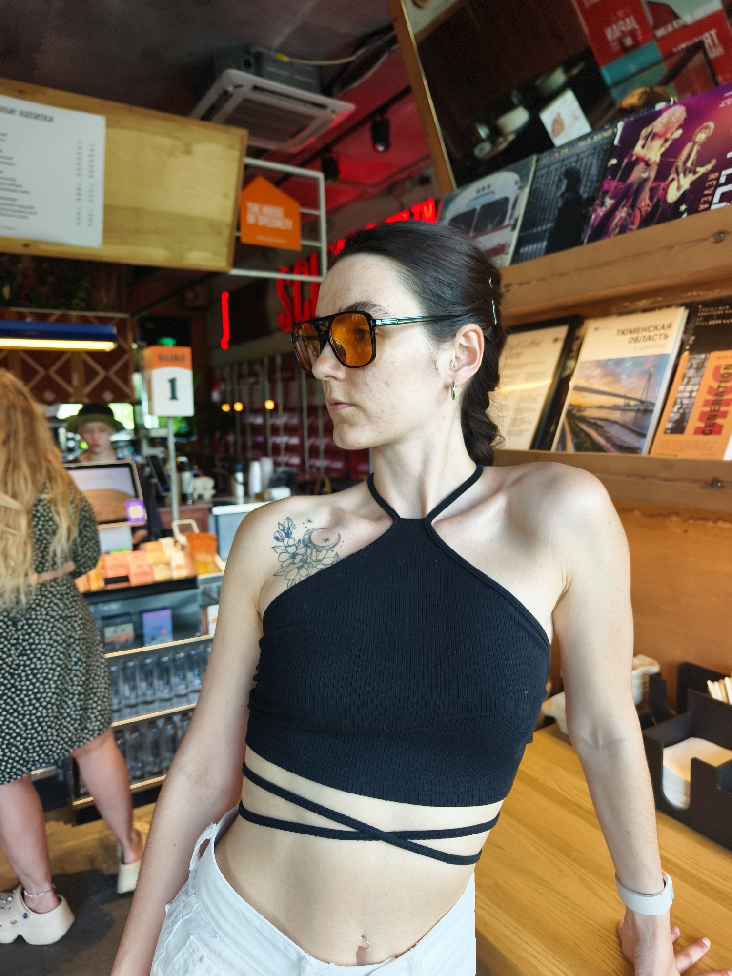 A medium shot captures a woman in a black crop top and white shorts, wearing orange sunglasses, inside a brightly lit establishment. The background features shelves filled with books and various items. To the left, another woman with blonde hair stands near a counter, and a person wearing a hat is visible behind the counter. A menu is displayed on a wooden board on the left wall, and a sign with the number "1" is visible above the counter. The environment appears to be a cafe or a similar public space. The city of Tyumen, Russia, is implied by the presence of books in Russian, such as "ТЮМЕНСКАЯ ОБЛАСТЬ" (Tyumen Oblast).