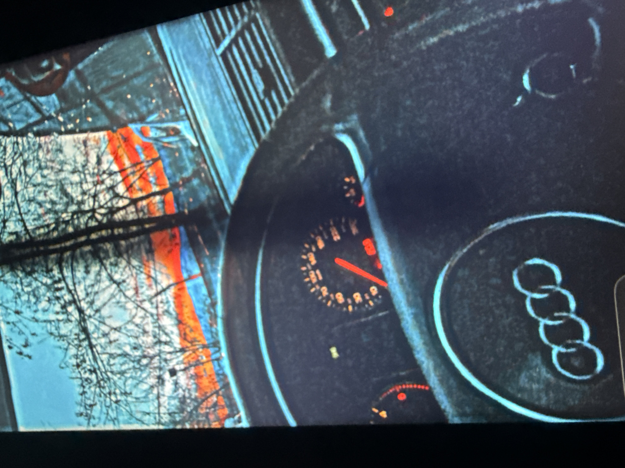 The image shows the interior of a car with a focus on the steering wheel and dashboard. Through the windshield, a sunset with a bright orange and red sky is visible, framed by bare tree branches. The dashboard features a speedometer with a red needle pointing towards approximately 60, and a digital display showing "205". The Audi logo is prominently displayed on the steering wheel. The scene appears to be captured from a stationary vehicle, possibly in a city like Sofia, Bulgaria, given the presence of urban elements in the background and the overall aesthetic often associated with European cityscapes. There are no people visible in the image. The lighting suggests it is either dusk or dawn.