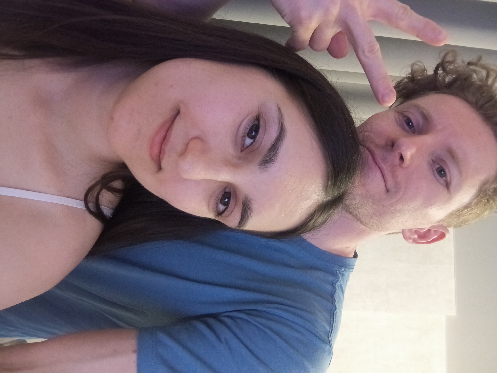 The image is a close-up, selfie-style shot of two people. In the foreground, a woman with long dark hair is looking directly at the camera with a slight smile. She is wearing a white strap. Behind her, a man with curly blonde hair and a beard is also looking at the camera and making a peace sign with his right hand. He is wearing a blue t-shirt. The background appears to be an indoor setting with a light-colored wall and some curtains. The scene is likely captured in a personal residence, potentially a bedroom or living area. There are no discernible landmarks or features that would allow for the specific identification of Krasnodar, Russia, as the location.
