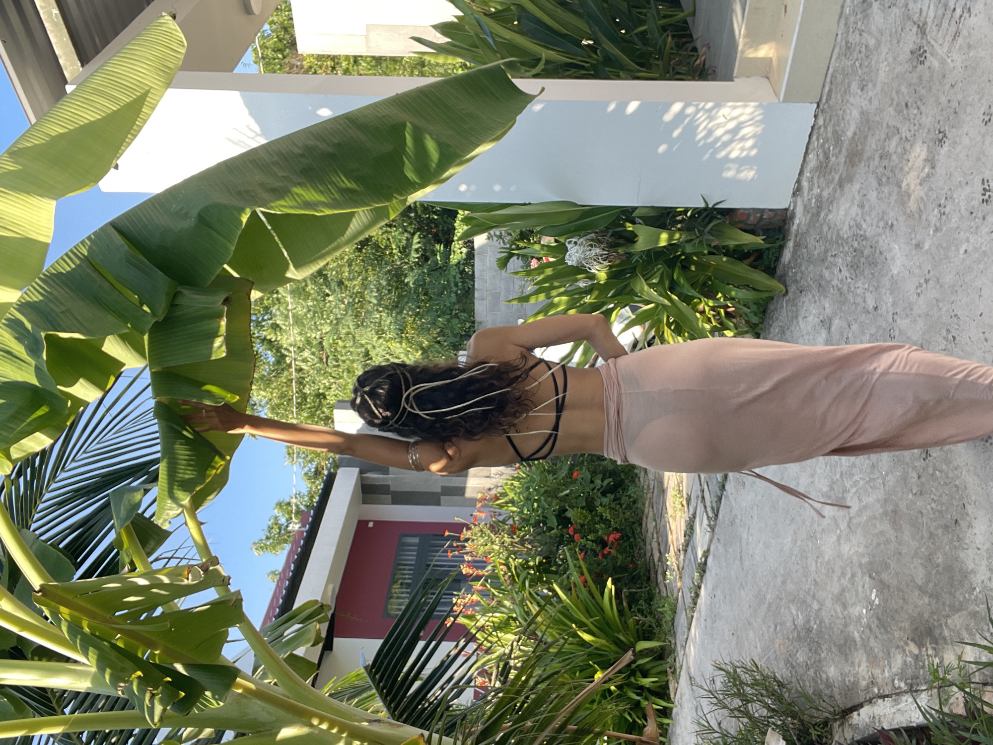 A woman stands with her back to the camera, reaching up to touch a large, green banana leaf. She is wearing a light pink, sheer skirt and a black bikini top. Her hair is curly and dark, adorned with a thin, metallic headband with white strings hanging down. She is standing on a concrete path in a garden setting. In the background, there is a red and white building with a gray roof, surrounded by lush green foliage and red flowers. The scene is bathed in bright sunlight. The location is described as Phu Thuy Ward, Vietnam, which is a rural area known for its agricultural land, including banana plantations.