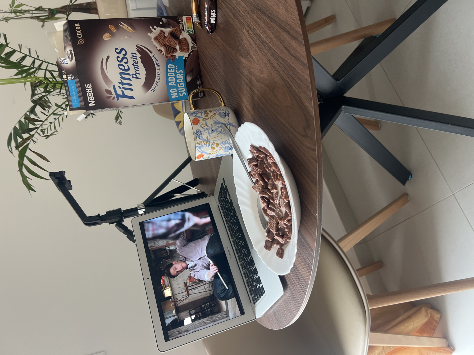 The image captures an indoor scene in Budva, Montenegro, focusing on a casual meal setup on a dark wooden table. A box of "Nestlé Fitness Protein" cereal, specifically "COCOA" flavor, is visible, highlighting "WHEAT, OATS & QUINOA" ingredients and "NO ADDED SUGARS." The box also displays "310g e," "NUTRI-SCORE ABCDE," and "CONTAINS MALTITOL," along with a manufacturing date of "01.2023" and other codes "L03770565 06:22 34." Nearby, a small "MINIS" branded wrapper is present.
A white ceramic bowl filled with chocolate-flavored cereal and milk, accompanied by a spoon, sits adjacent to an open MacBook Air laptop. The laptop screen displays a video, showing a man in what appears to be a rustic or workshop-like interior, holding a long stick or tool. A decorative white ceramic mug with blue, orange, and gold floral patterns and a gold handle is placed between the cereal box and the bowl.
A black tripod or stand is positioned to the left of the laptop. In the background, a green houseplant adds a touch of natural greenery. The floor is covered with light-colored tiles, and parts of a chair with wooden and black metal legs are visible. The overall scene suggests an individual enjoying cereal while watching content on a laptop, likely during a relaxed moment indoors.