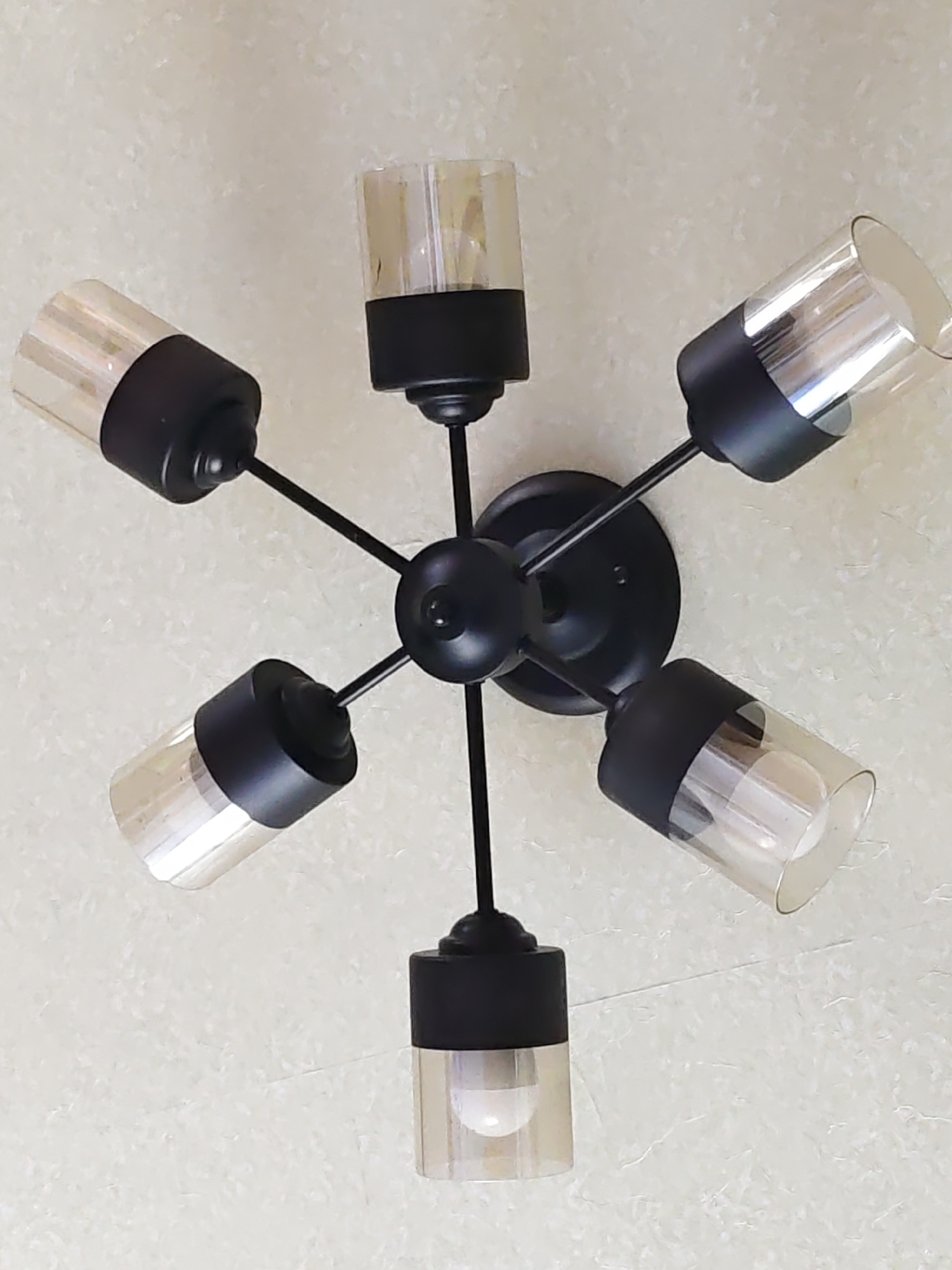 The image shows a close-up, overhead view of a black, multi-arm ceiling light fixture mounted on a light-colored, textured ceiling, likely in Rostov-on-Don, Russia. The fixture has five arms extending from a central hub, each supporting a black socket with a cylindrical glass shade. The glass shades are clear with a subtle amber tint and contain visible light bulbs. The fixture is centrally positioned in the frame, with the ceiling occupying the majority of the background.