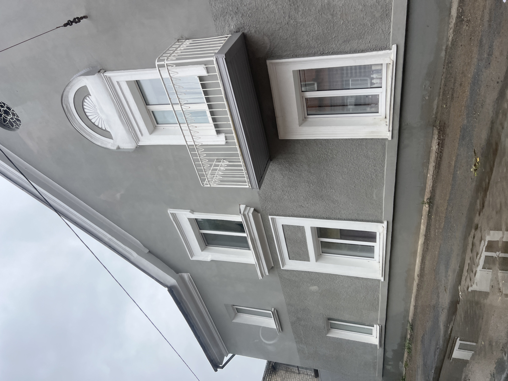 The image shows the exterior of a two-story building with grey stucco walls and white trim around the windows and along the roofline. There are several windows of various sizes on the facade. A small balcony with a white railing juts out from the upper story. Above the balcony, there is an arched decorative element with a fan-like pattern. A circular decorative grille is visible higher up on the facade. The building is situated on a street with puddles of water on the asphalt, reflecting the building. The sky is overcast. The building appears to be located in the city of Ulyanovsk, Russia.