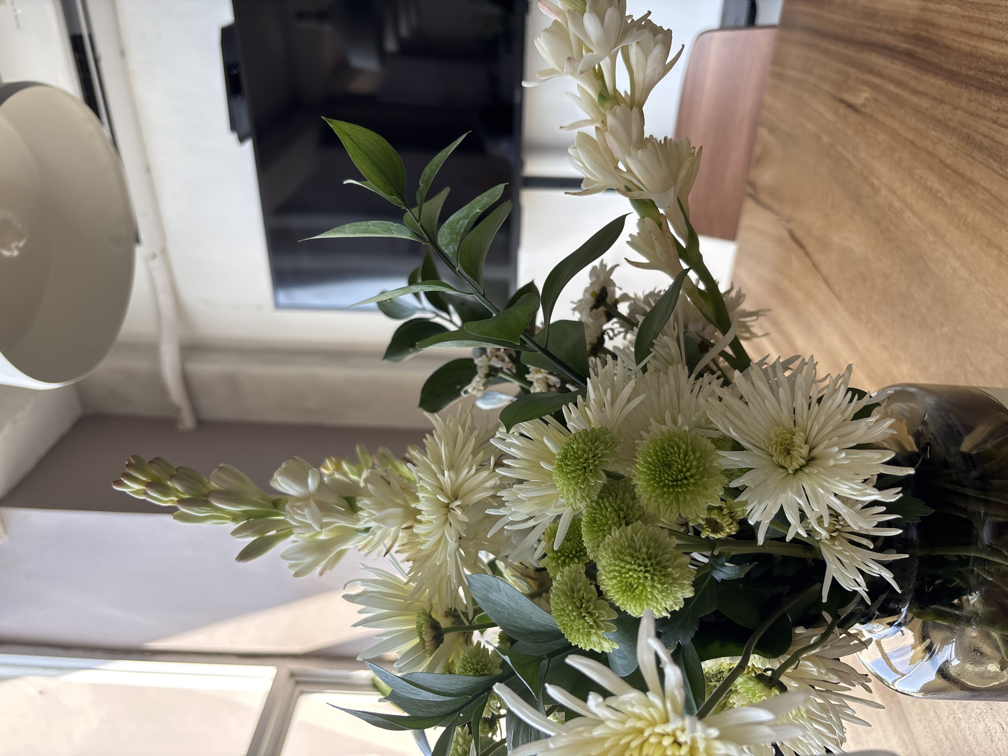 A close-up, eye-level shot showcases a bouquet of white and green flowers in a clear glass vase. The flowers are arranged on a wooden table, with a window and a television in the background. The sunlight streams in from the left, illuminating the flowers and creating soft shadows. The scene appears to be indoors, in an area that could be a home or office. The Kuta Selatan area of Indonesia is known for its beaches and resorts, but this image depicts an interior space, possibly a restaurant or hotel lounge, where the bouquet might be a decorative element.
