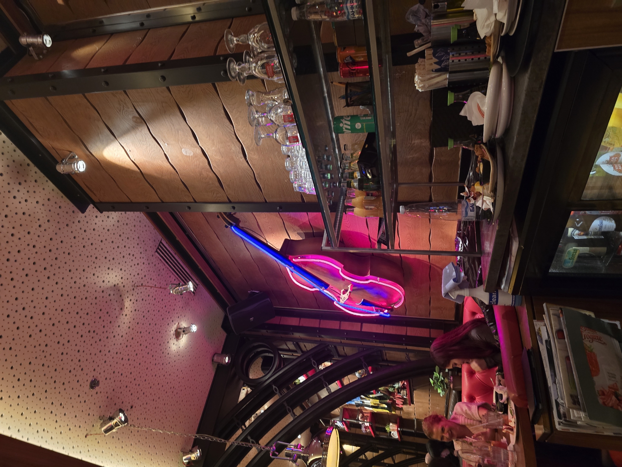 The image shows the interior of a bar or restaurant. The perspective is from an unusual angle, looking upwards. The ceiling features a wooden design with black metal supports and recessed lighting. A neon sign in the shape of a violin hangs from the structure. Shelves are stocked with glassware, bottles of alcohol, and assorted bar supplies. The wall to the left has a textured surface with numerous small holes and spotlights. Below, the lower portion of the scene displays a bar area with people seated at a counter. There are red chairs, menus, and other miscellaneous items. The details of the city are not discernible from the image. Sofia, Bulgaria, is only visible through deduction of the setting.