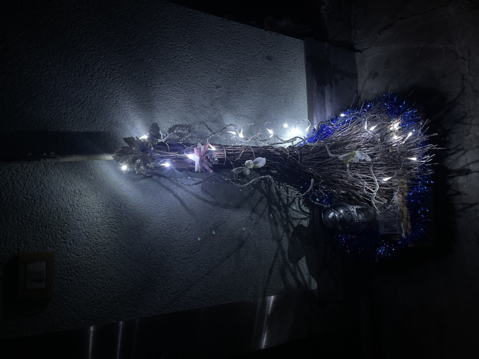 The image displays a decorated broom, positioned horizontally against a textured wall in what appears to be a dark indoor setting. The broom, likely a decorative piece or "witch's broom," is made of bundled twigs and branches. It is elaborately adorned with numerous cool white LED string lights that brightly illuminate the immediate area and cast shadows on the wall behind it.
Towards the left, where the handle would be, the broom features silver-colored decorative leaves and a small red and white checkered bow or star. Further along the bundled branches, more white or light-colored floral decorations are visible. The rightmost end of the broom is embellished with bright blue tinsel, adding a vibrant color contrast to the otherwise dark and muted tones of the scene. Small metal objects, possibly bells or part of the decoration, can be seen nestled within the branches on the right.
The wall behind the broom has a rough, textured finish, painted in a dark gray or blue-gray hue. In the bottom left corner of the image, an electrical light switch is visible on the wall, partially illuminated by the lights from the broom. The overall scene is dark, suggesting it is nighttime or a dimly lit room, with the string lights being the primary source of illumination. The strong artificial light creates a festive and somewhat magical atmosphere. There is no visible text in the image. The setting is in Castione, Italy.