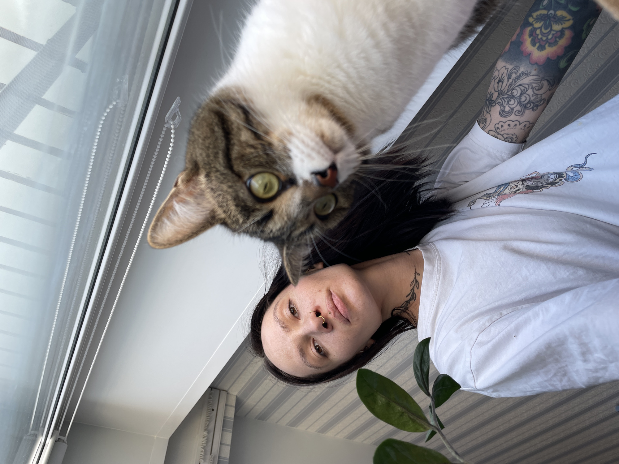 The scene is set indoors within a residential environment in Izhevsk, Russia. A low-angle perspective captures an adult female and a domestic cat in the foreground. The female, positioned on the left and center, has long dark hair and an unsmiling expression, looking directly towards the camera. She has visible facial piercings, including a gold-colored septum ring, a stud on her right nostril, and a labret piercing below her lower lip. A leafy vine tattoo is visible on the right side of her neck. She wears a white short-sleeved t-shirt, revealing a colorful graphic design on the left side featuring a skull and a snake, and a full sleeve tattoo on her left arm comprising intricate floral and paisley patterns with a prominent colorful flower on a darker background. To the right and in the foreground, a domestic cat with brown and white tabby markings and bright green eyes looks towards the camera. The cat's head is slightly out of focus due to its close proximity. The background on the left features light grey and white striped wallpaper and green leaves of an indoor plant. The upper right portion of the image shows a white window frame with integrated roller blinds and bead chains, reflecting a bright, indistinct outdoor sky.